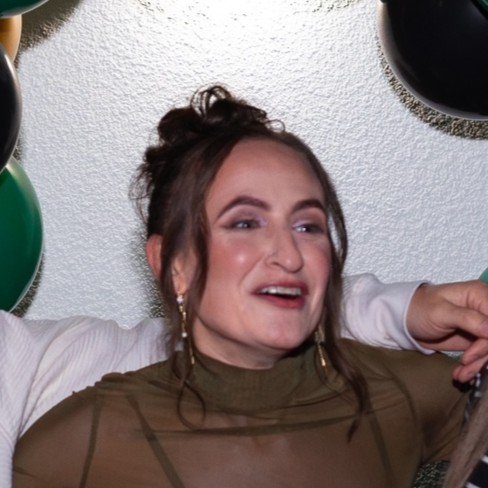A woman with brown hair in a messy bun, wearing earrings and a brown top, smiling and looking to the side, with her arms raised and balloons around her.