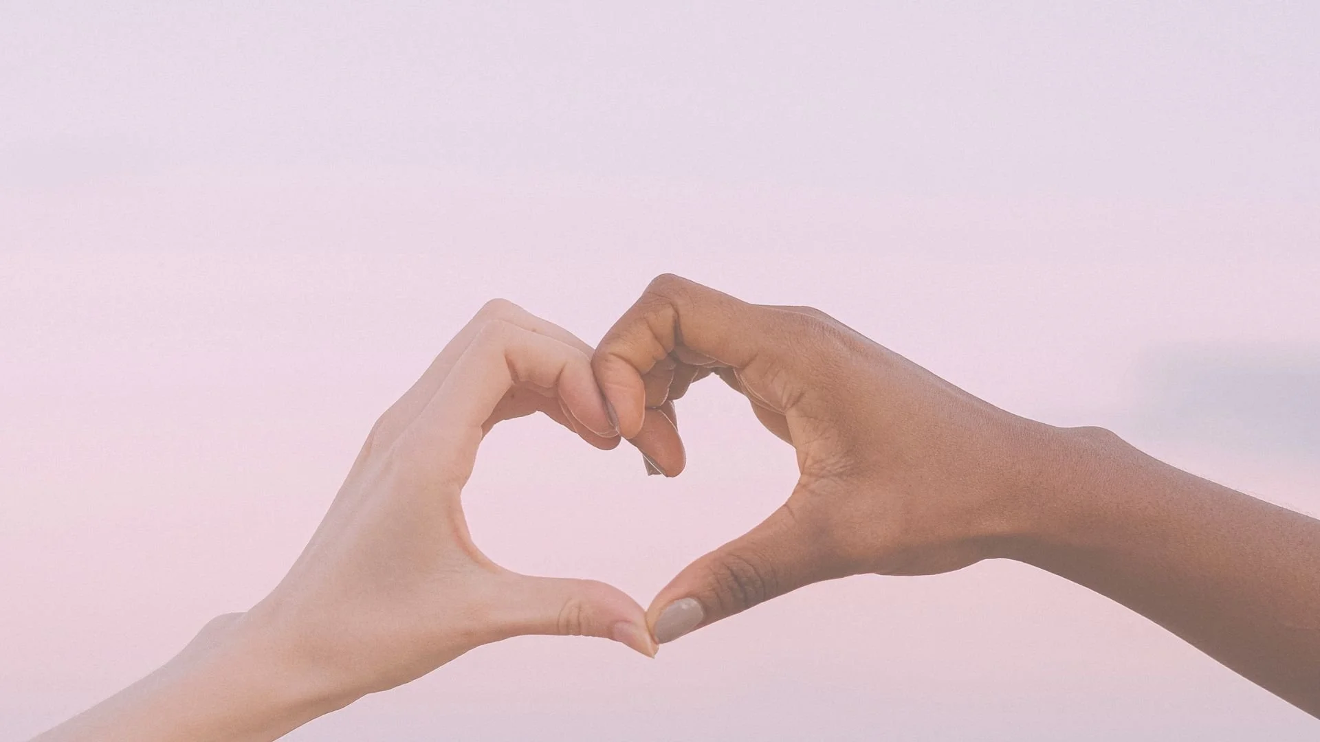 Two hands, one light-skinned and one dark-skinned, forming a heart shape against a pale pink background.