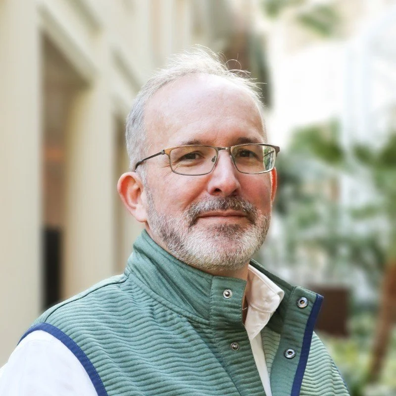 A middle-aged man with glasses, short gray hair, and a beard, wearing a green quilted vest over a white shirt, standing outdoors with blurred greenery and buildings in the background.