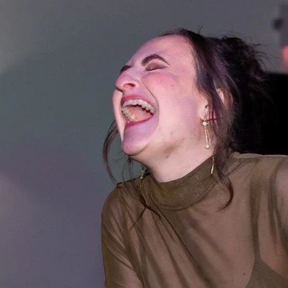 A woman laughing joyfully with her head tilted back, wearing a sheer brown top and gold earrings.