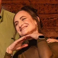 A woman smiling and resting her chin on her hand, sitting indoors with a wooden background.