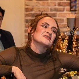 A woman with reddish-brown hair, wearing a black long-sleeve top, making a playful facial expression, in an indoor setting with a brick wall background.