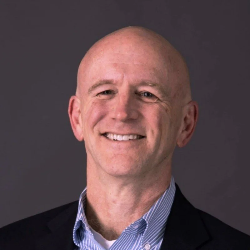 A smiling bald man wearing a black blazer and a light blue collared shirt against a dark background.