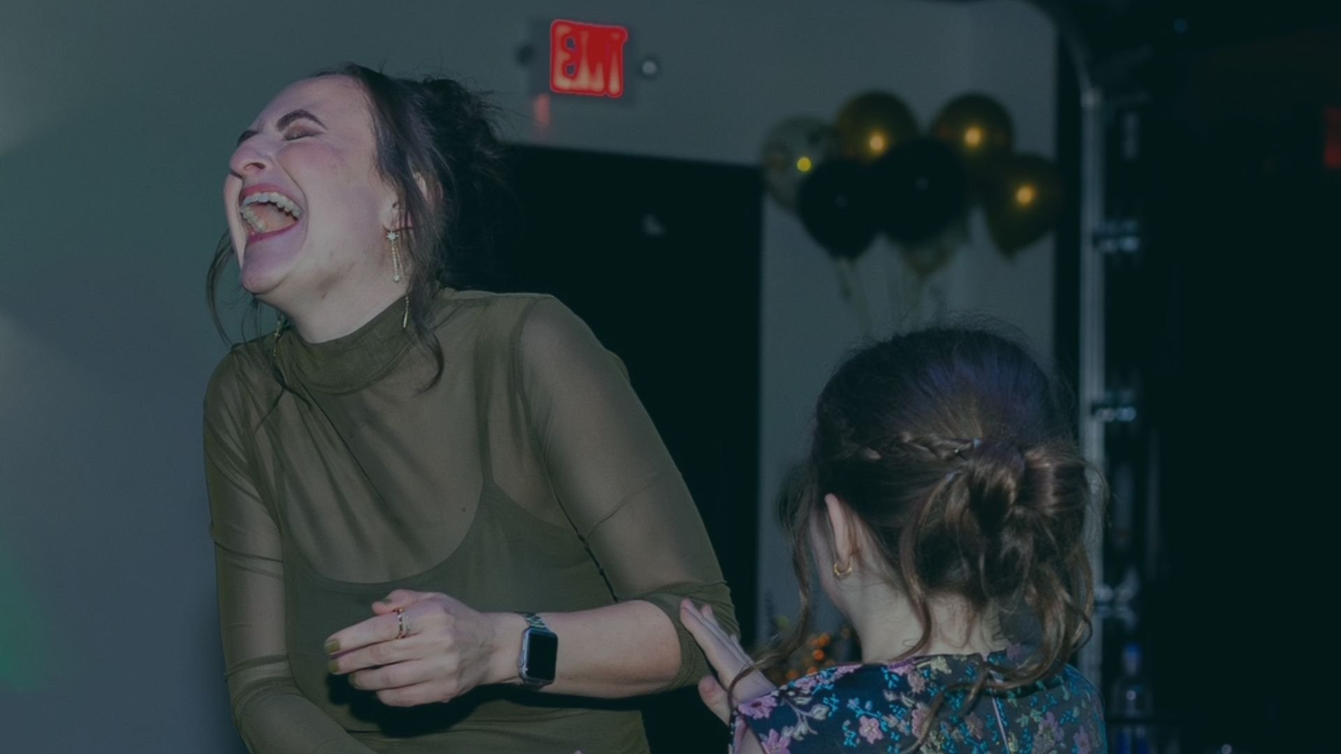 Two women laughing and smiling in a room with black and gold balloons in the background.