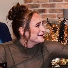 Profile of a smiling woman with red hair styled in an updo, wearing a sheer long-sleeve top, sitting indoors with a brick wall and decorative plants in the background.