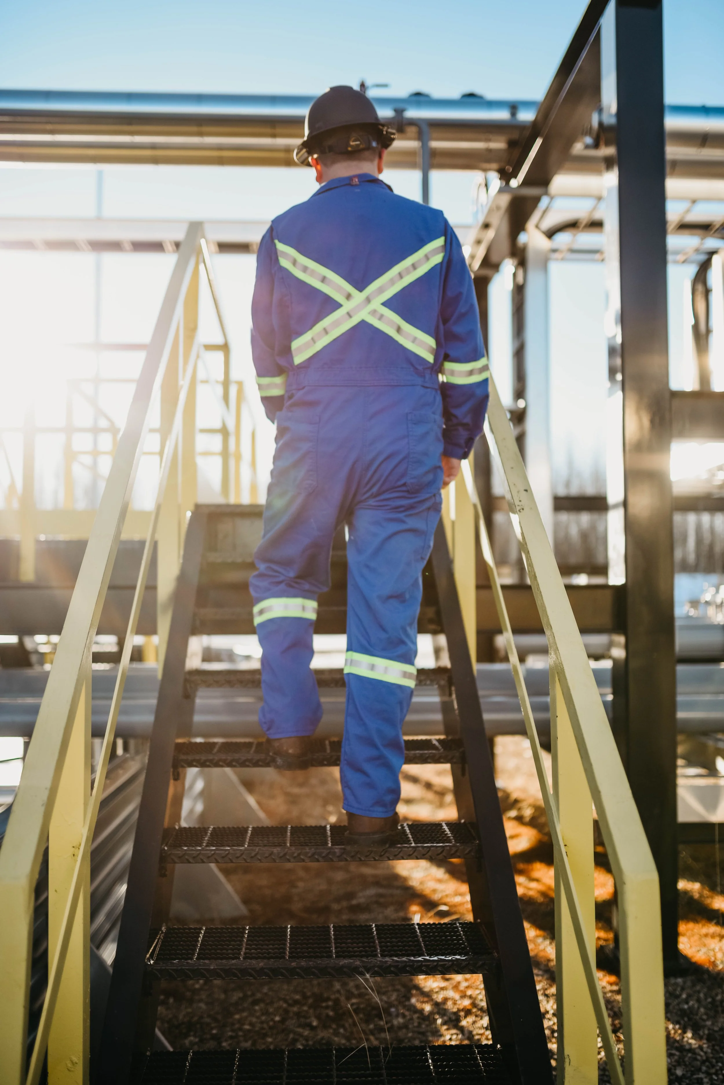 A worker in blue coveralls and a safety helmet climbing metal stairs on an industrial site, with pipes and equipment visible in the background.