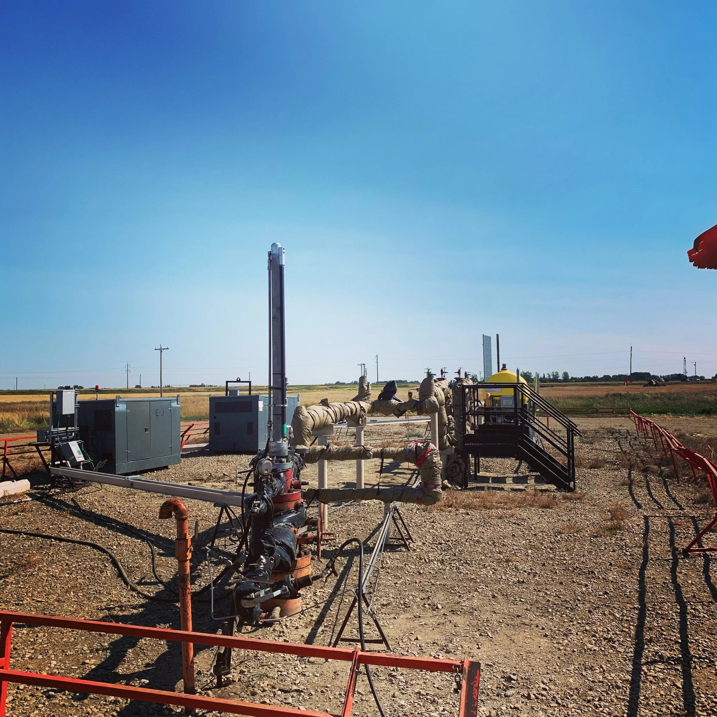 Oil or gas extraction equipment in a rural field under clear blue sky, with pipelines and machinery.