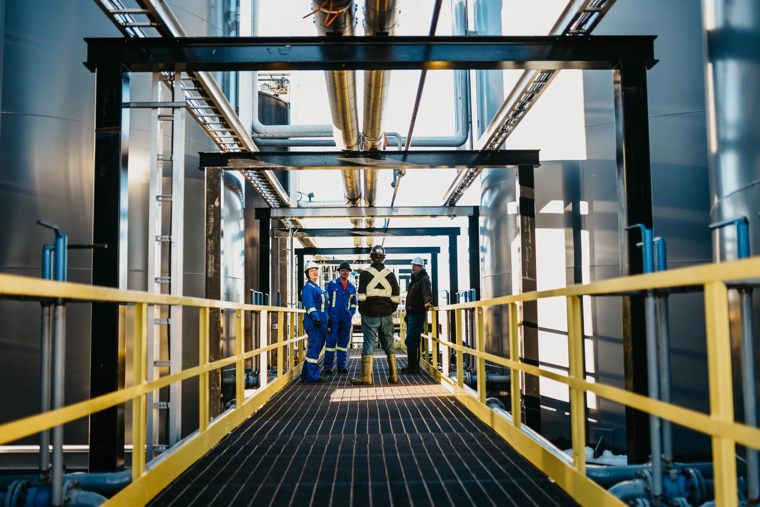 Four workers in safety gear, including helmets and reflective clothing, standing and talking on a catwalk inside an industrial facility with pipes and metal structures.