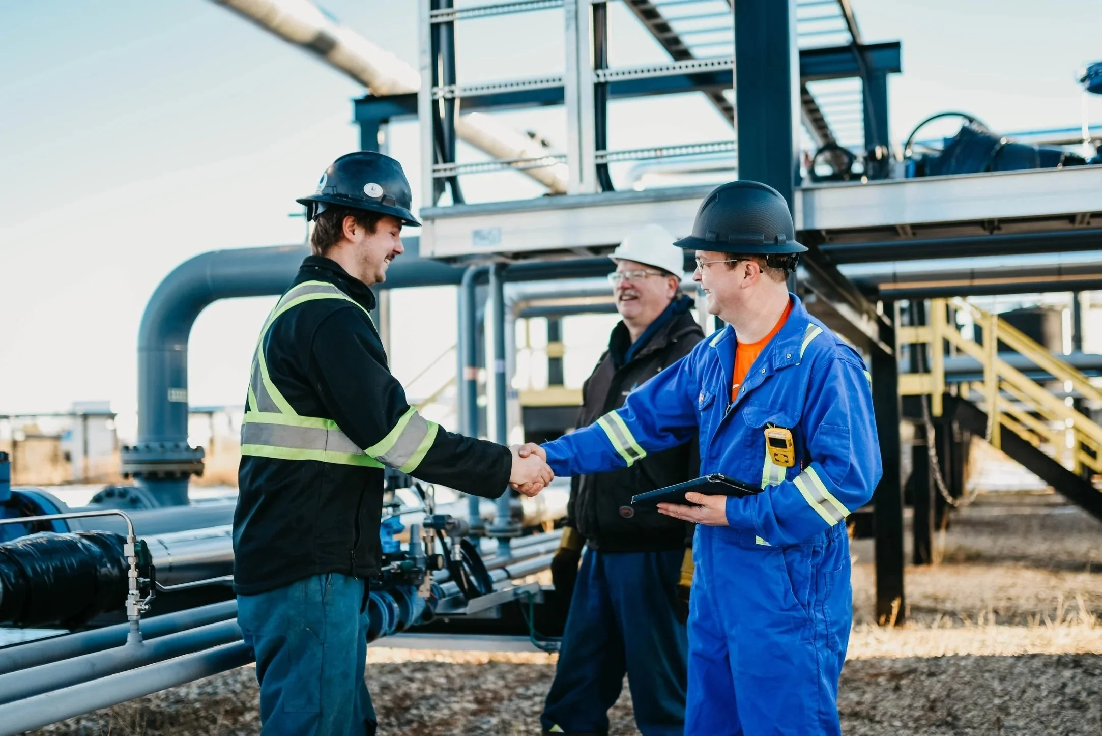 Three industrial workers in safety gear shaking hands at an outdoor industrial site, with pipes and metal structures in the background.