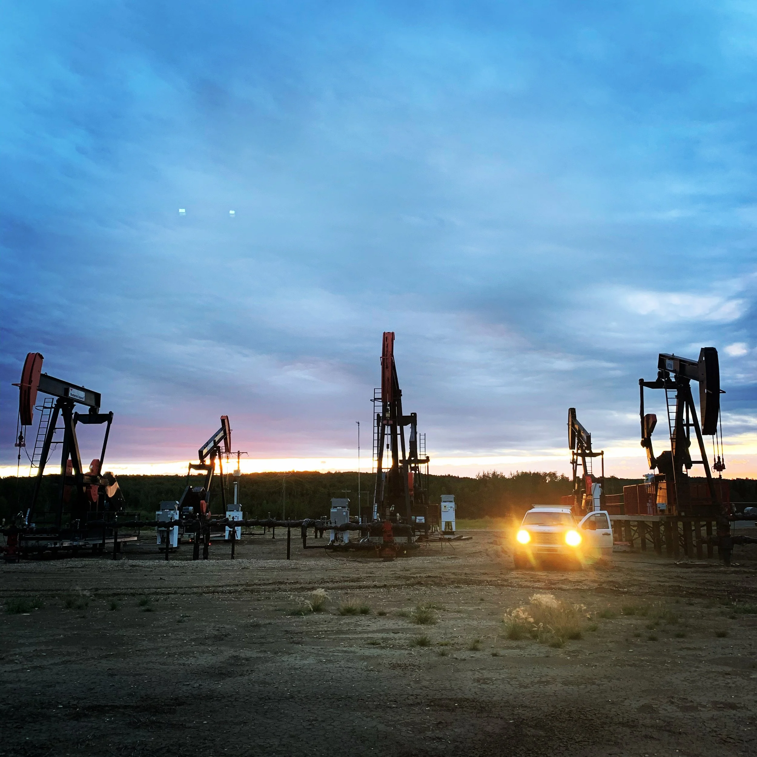 Oil pumpjacks operating at sunset on a flat dirt field with a barely visible car with headlights on in the foreground.