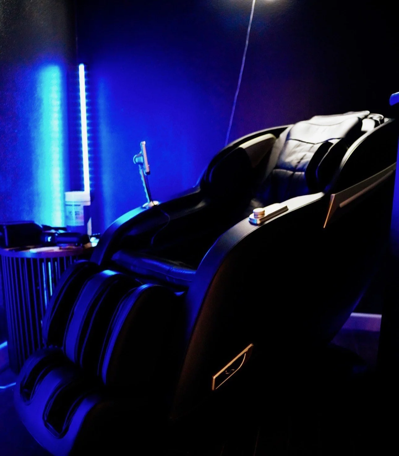 A massage chair with black leather upholstery in a dimly lit room with blue LED lighting.