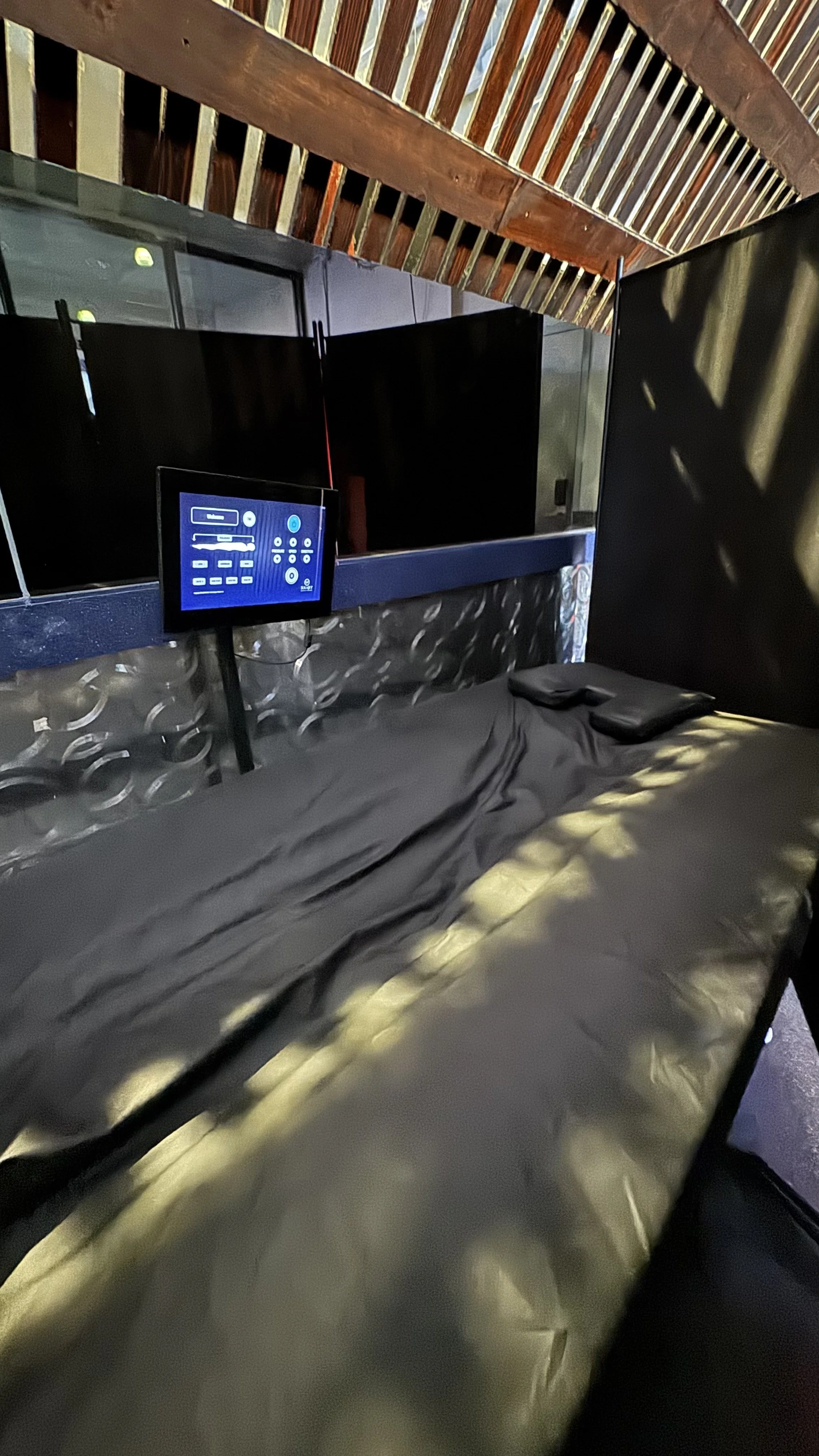 A black bed with two black pillows and a black bedsheet, in a room with wooden beams on the ceiling and a metallic wall. A small TV or monitor is mounted on a stand next to the bed.