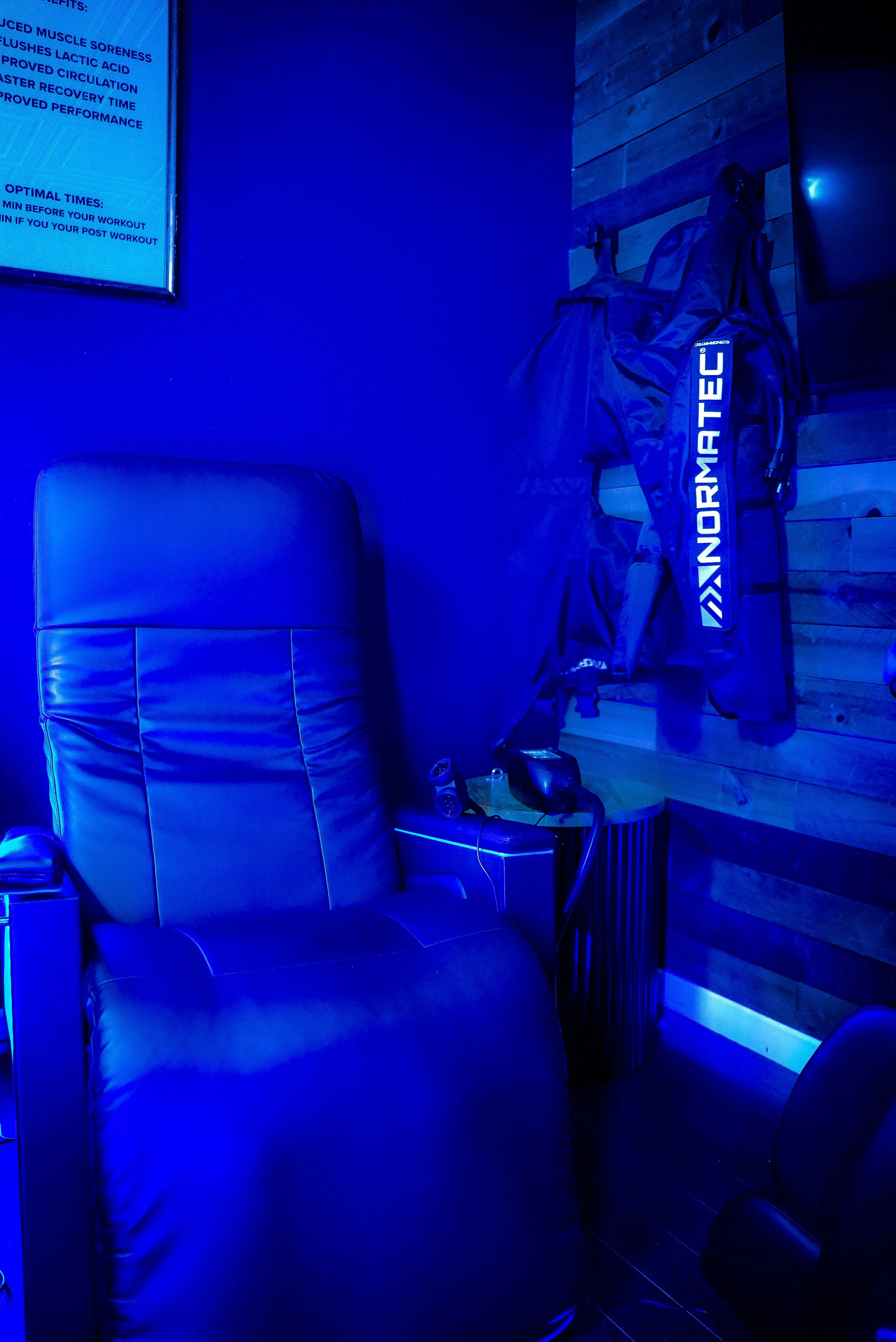 A room illuminated in blue light, featuring a leather recliner, a side table with a telephone and a water bottle, and gym clothing and a bag hanging on the wooden wall.