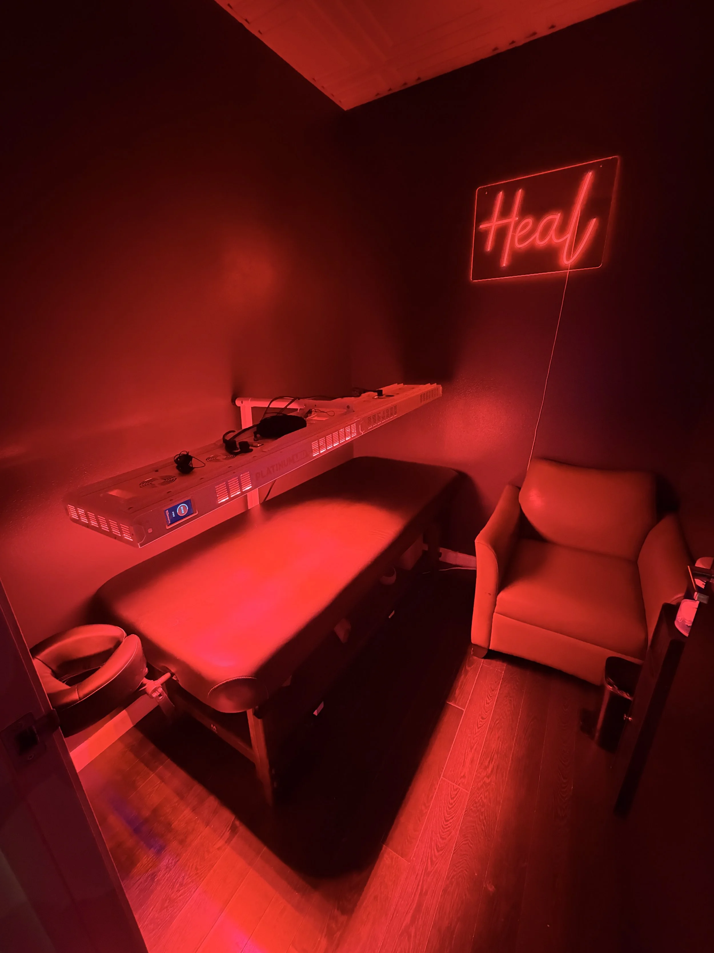 A dimly lit room with red neon lighting, a massage table, a neon sign that says "Heal," and a cozy armchair.