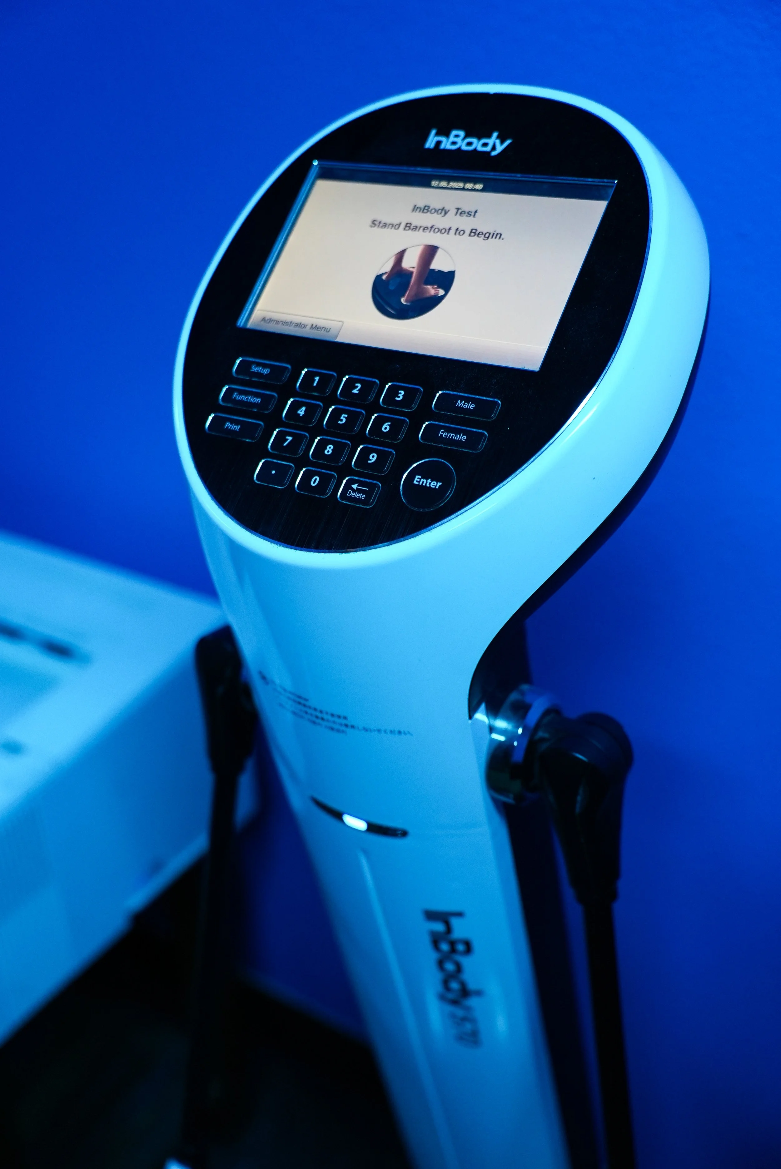An InBody body composition analyzer machine with a digital screen displaying a test prompt to stand barefoot to begin, in a room with blue lighting.