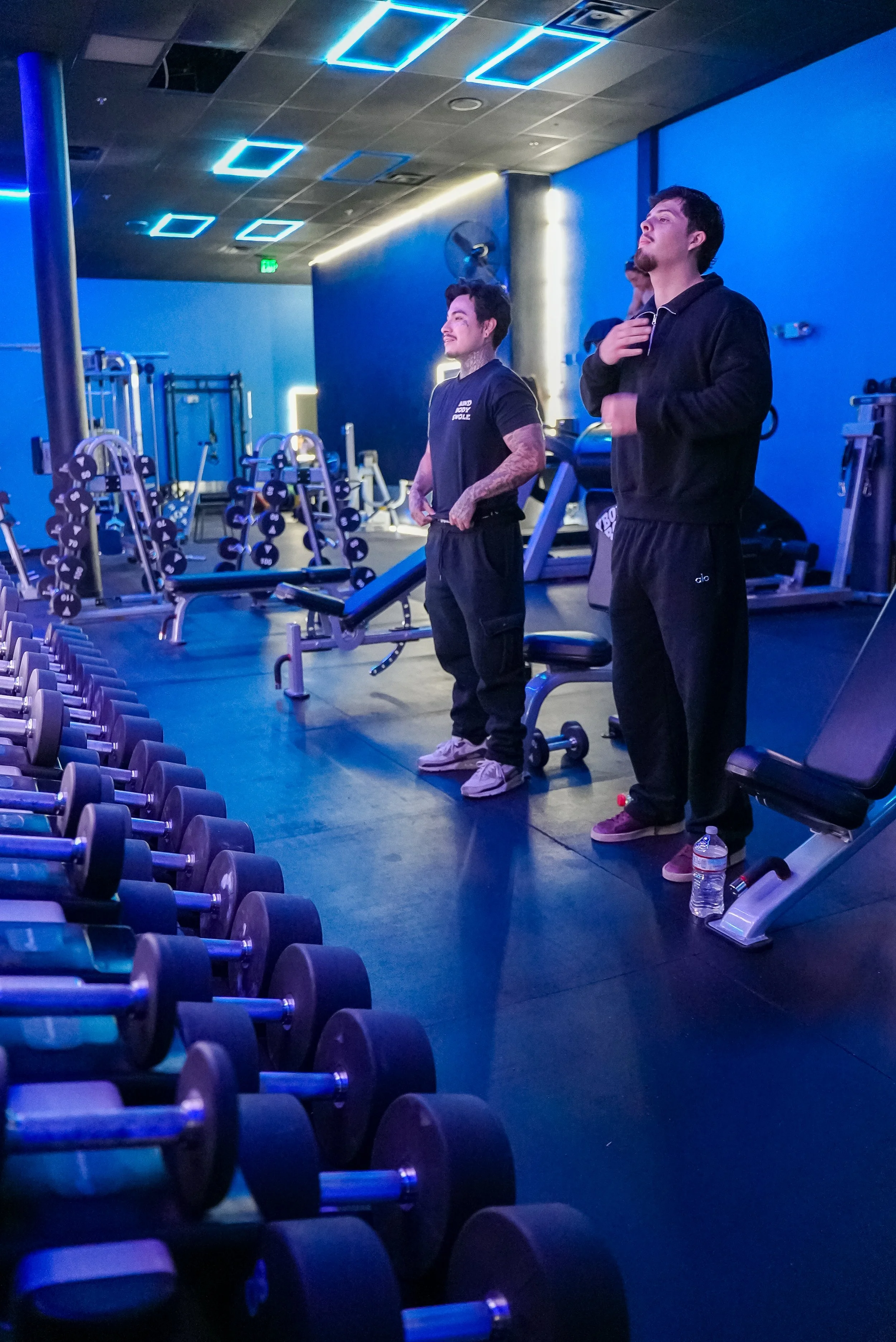 Three people standing in a gym with blue lighting, surrounded by various gym equipment including dumbbells, lift machines, and benches.