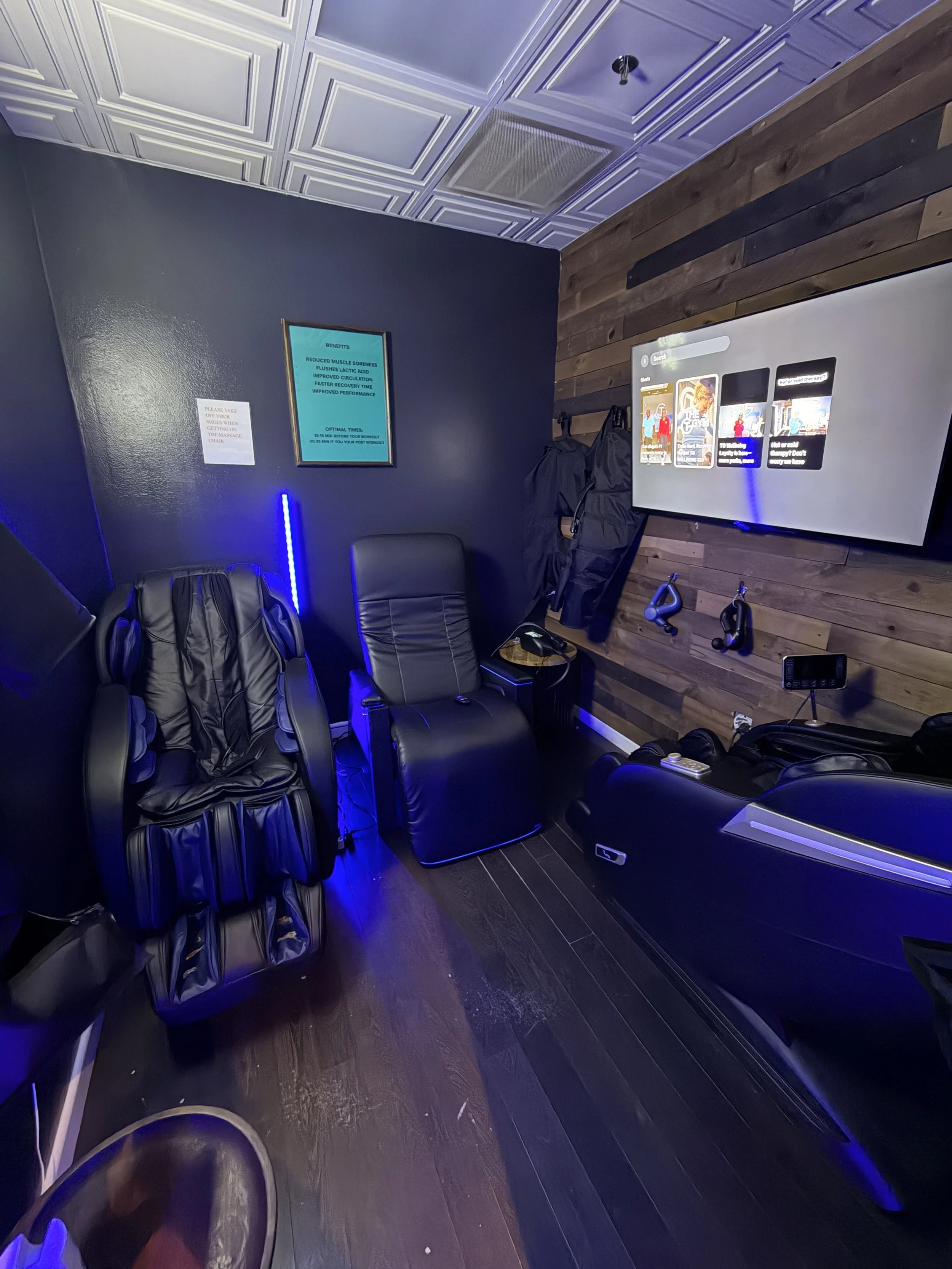 Room with massage chairs, a wall-mounted TV, and massage equipment, illuminated with blue lighting accents.