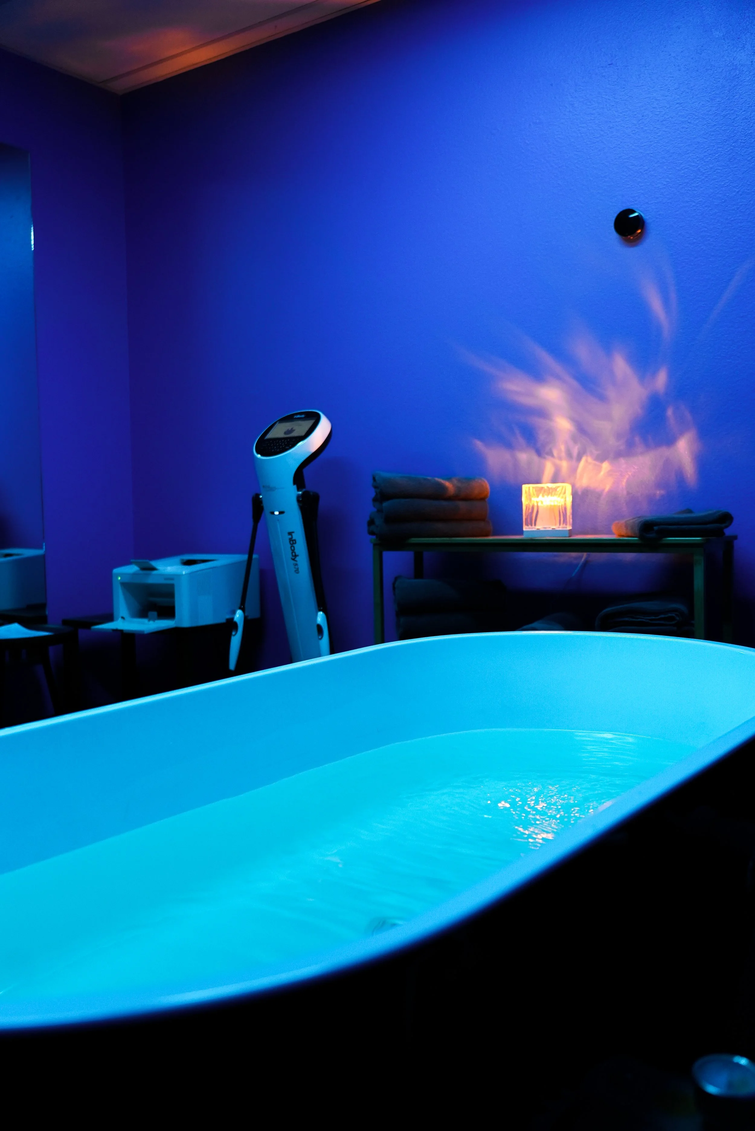 A spa room with a blue-lit bathtub, towels on shelves, a candle, and a body analysis machine.