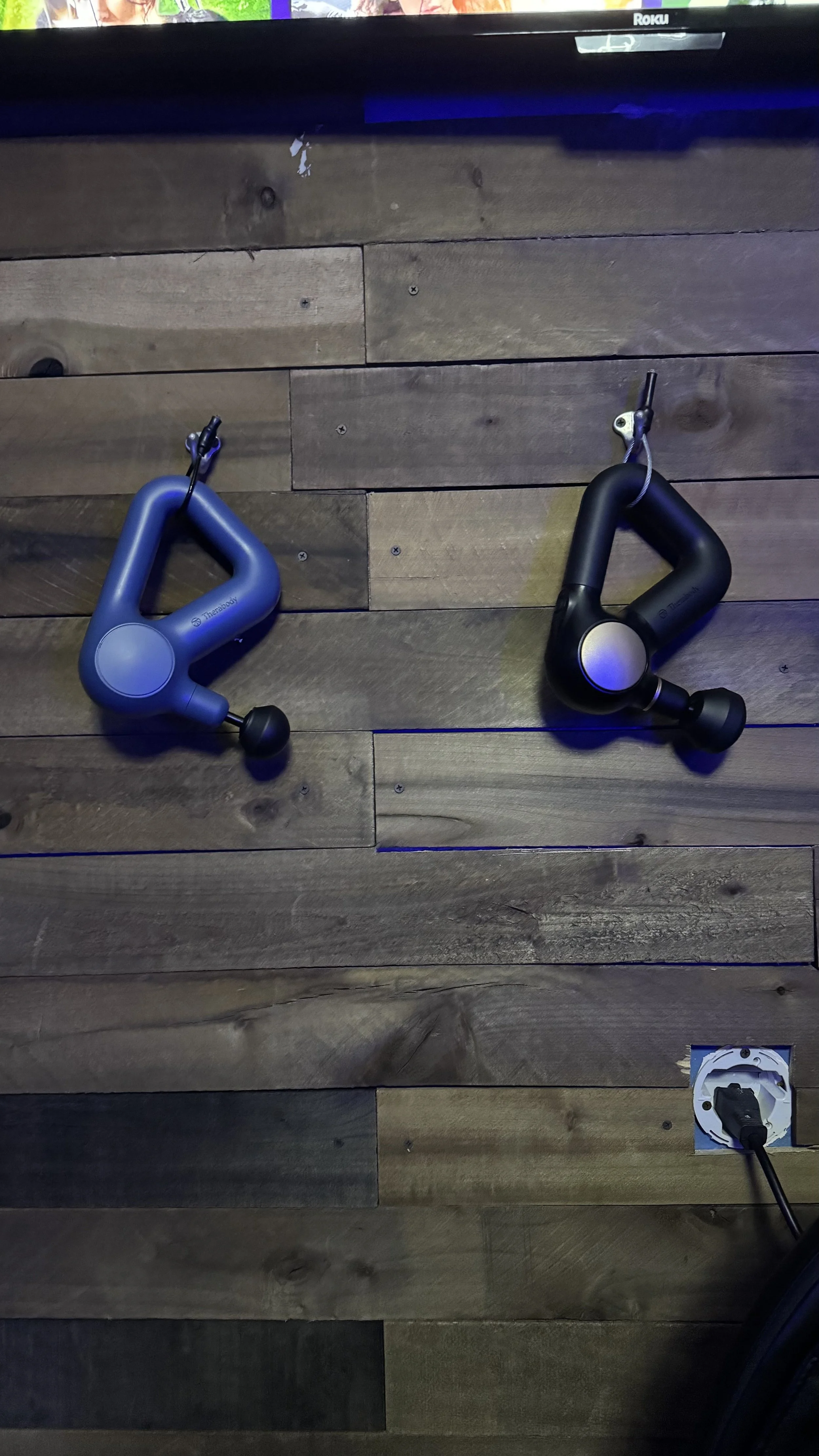 Two black and blue Theragun massage devices mounted on a wooden wall, with a power outlet and cord visible at the bottom right.