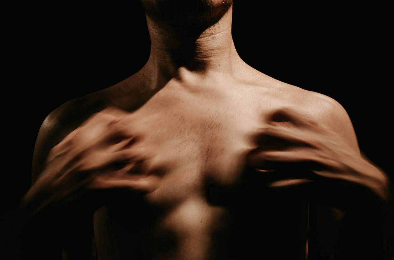 A shirtless man adjusting his chest with his hands against a dark background.