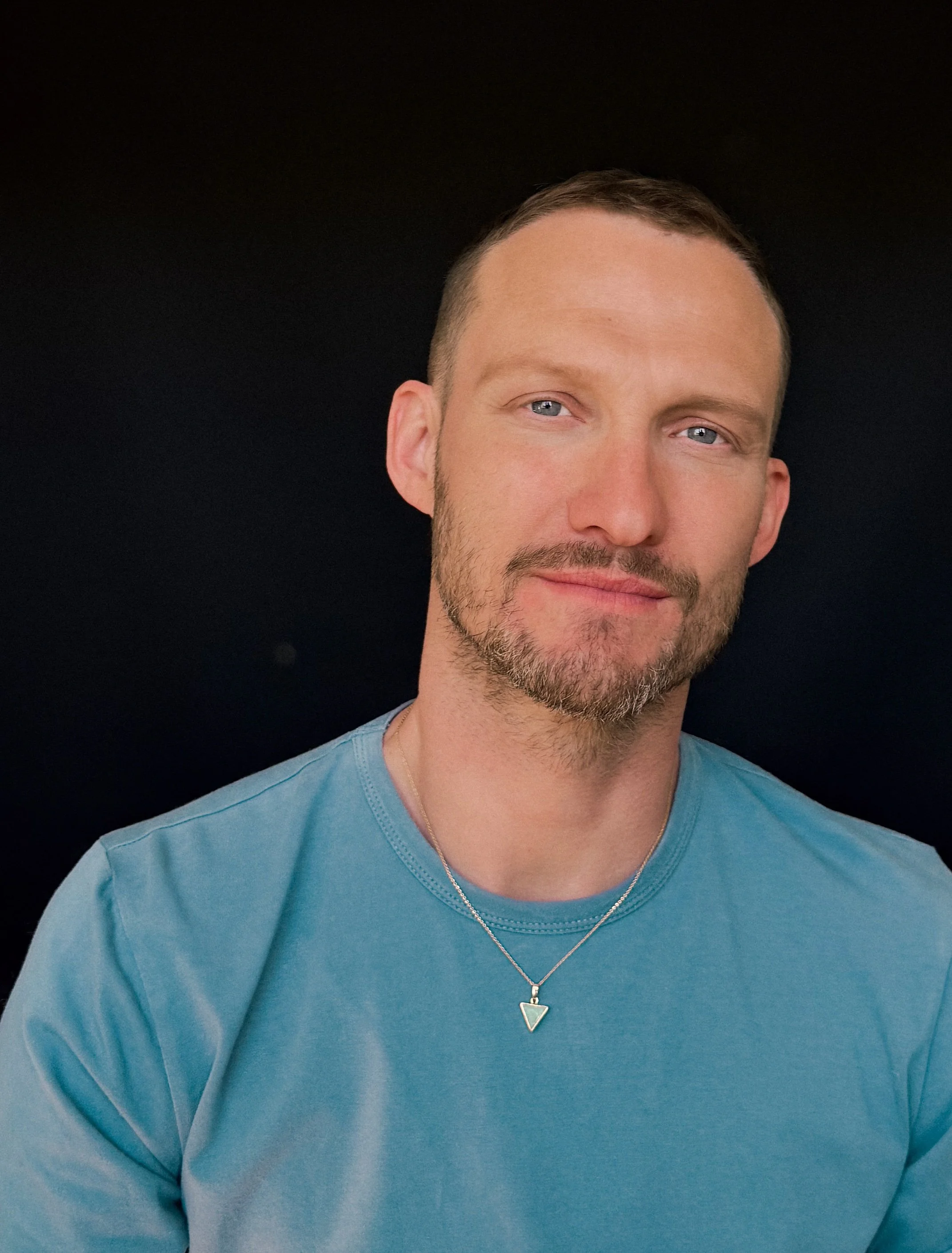 Head and shoulders portrait of a man (sex therapist for gay men Gary Albert) with short hair, light blue eyes, light complexion, beard, wearing a light blue t-shirt and a silver necklace with a small triangular pendant, against a black background.