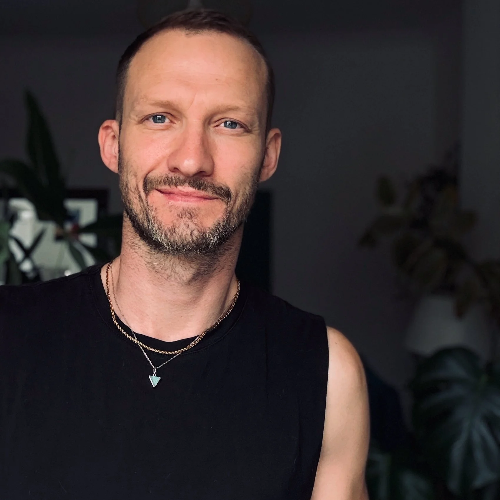 Close-up of a smiling man with short hair, beard, and light blue eyes wearing a black tank top, necklaces, and a gray baseball cap, with blurred dark background and some greenery.
