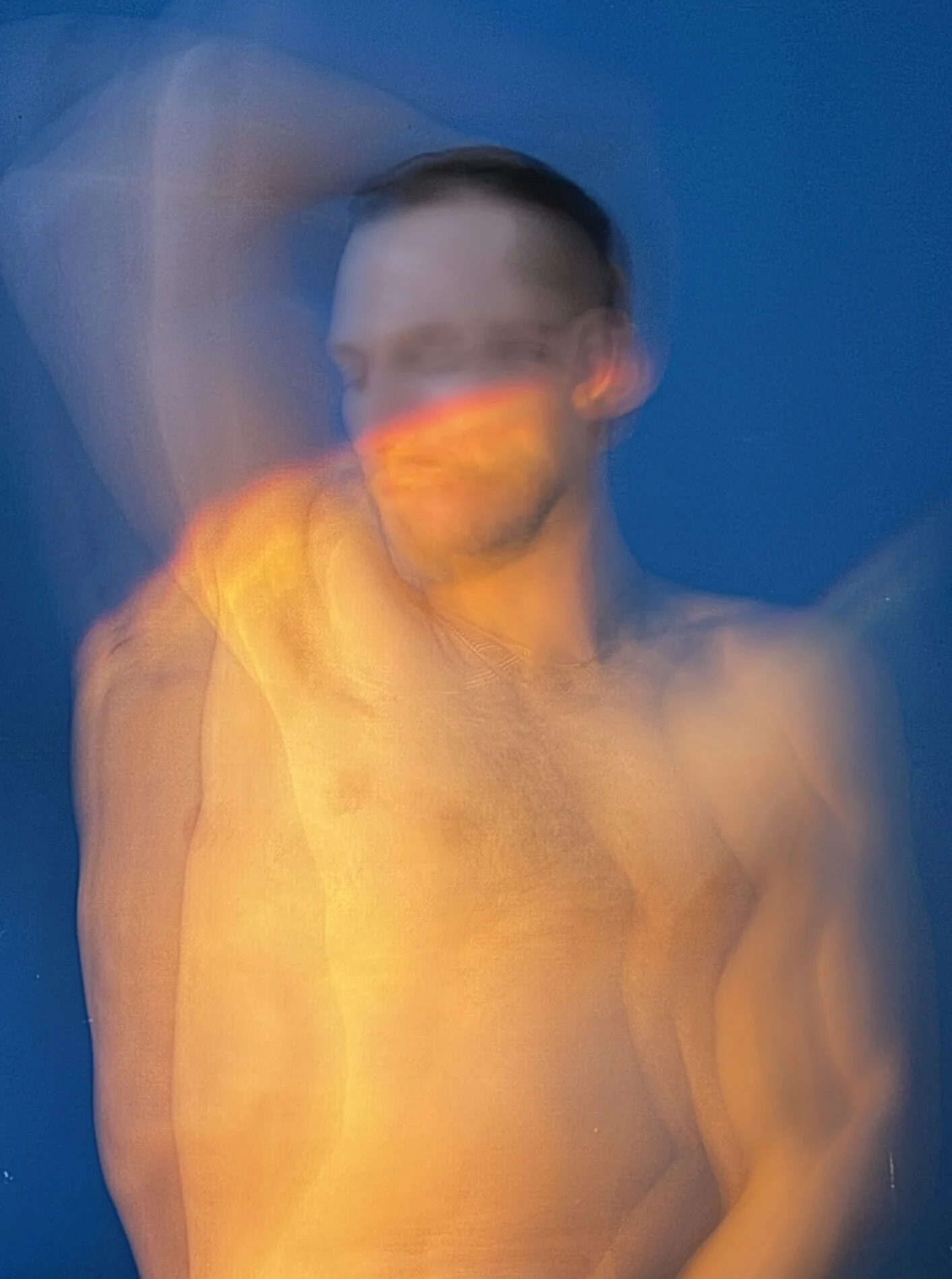 A man with short dark hair and a beard, shirtless, stands against a blue background with a blurred motion effect, creating a sense of movement.