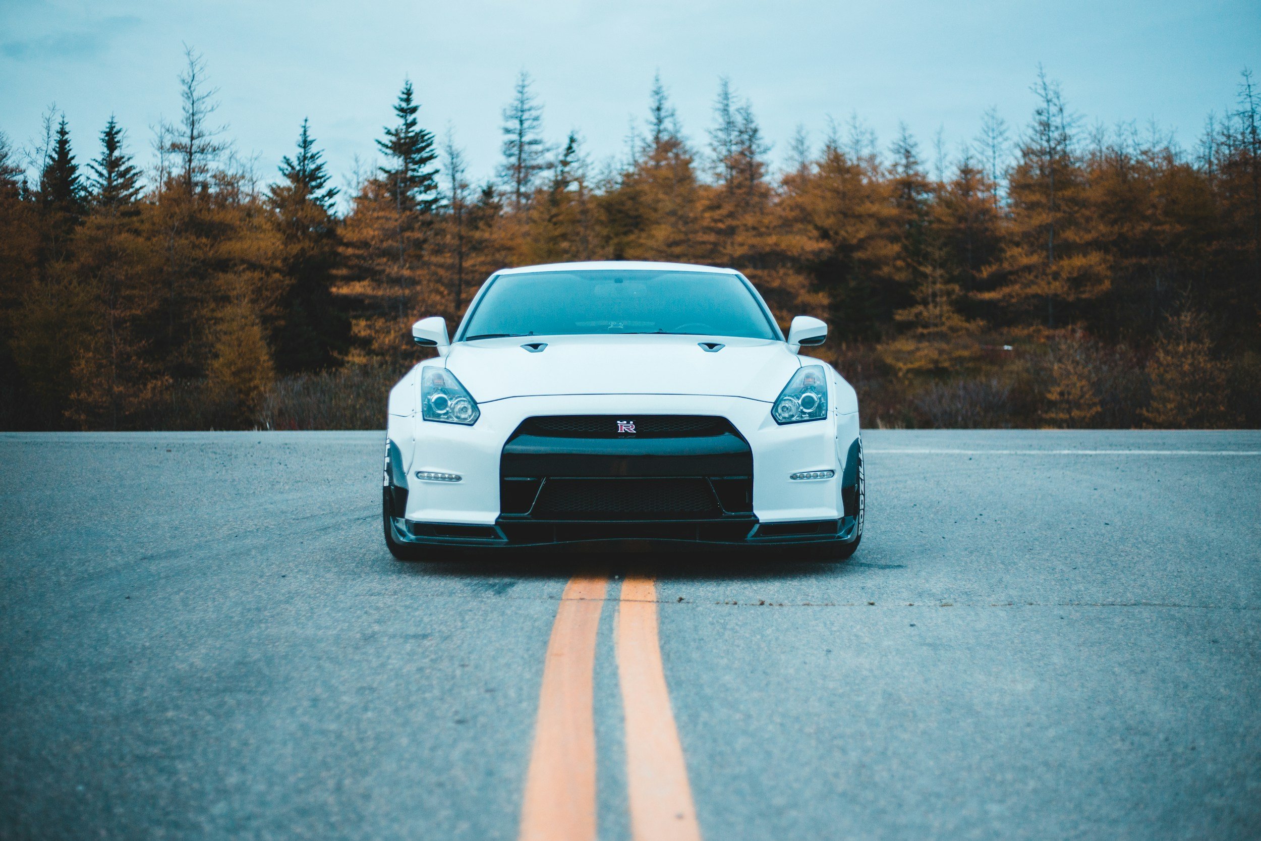 White Nissan GT-R sports car parked on an empty road with double yellow lines, and autumn trees with colorful leaves in the background.