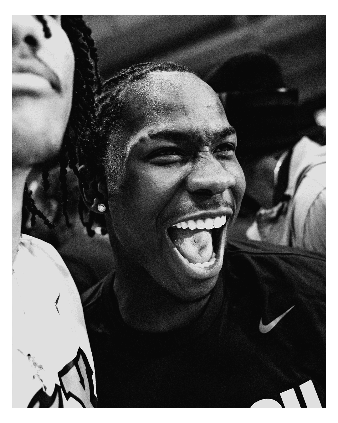 Close-up of a person with dark skin, short curly hair, smiling broadly with mouth open, showing teeth, wearing a dark shirt and earrings, in a black and white photo.