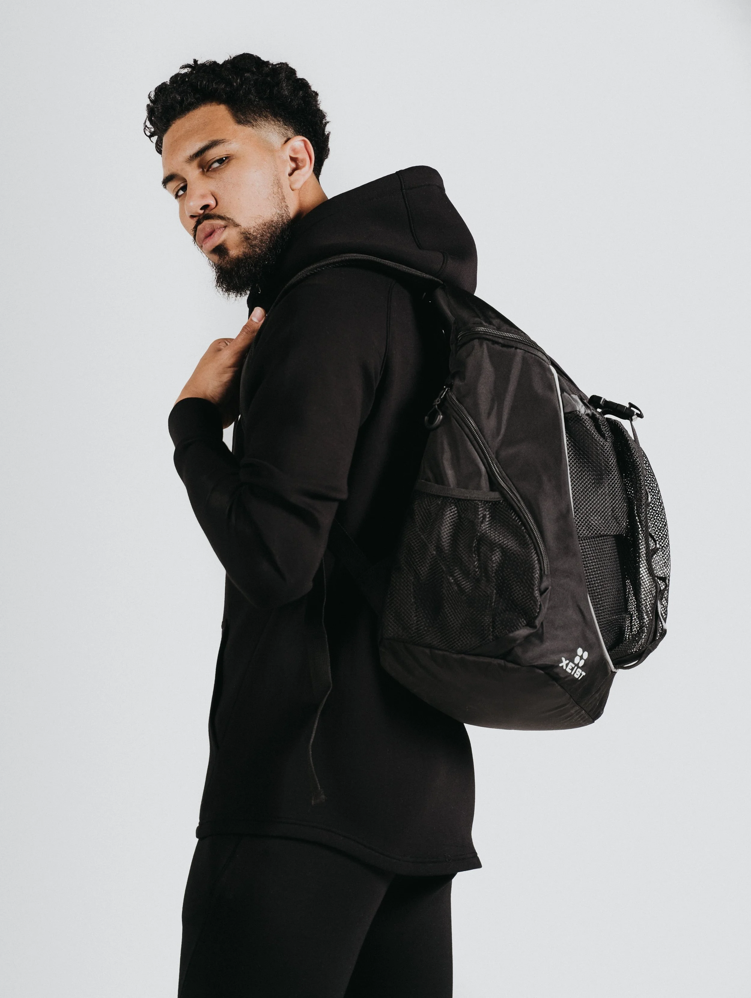 Young man with curly hair and a beard wearing a black hoodie and carrying a black backpack, looking over his shoulder against a plain white background.