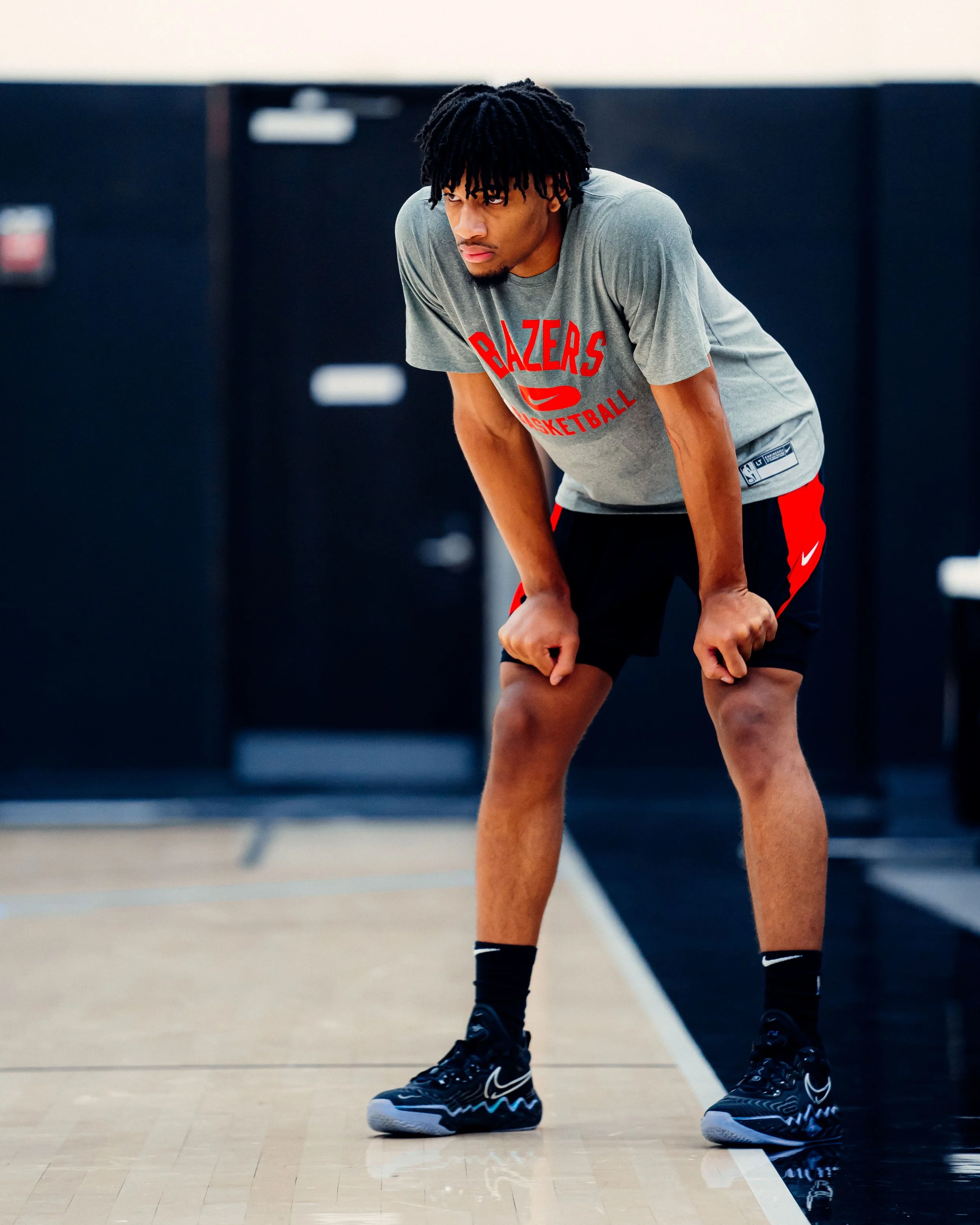 A young man with dreadlocks wearing a gray Portland Trail Blazers basketball t-shirt and black shorts, bent over with hands on knees, practicing on a basketball court.