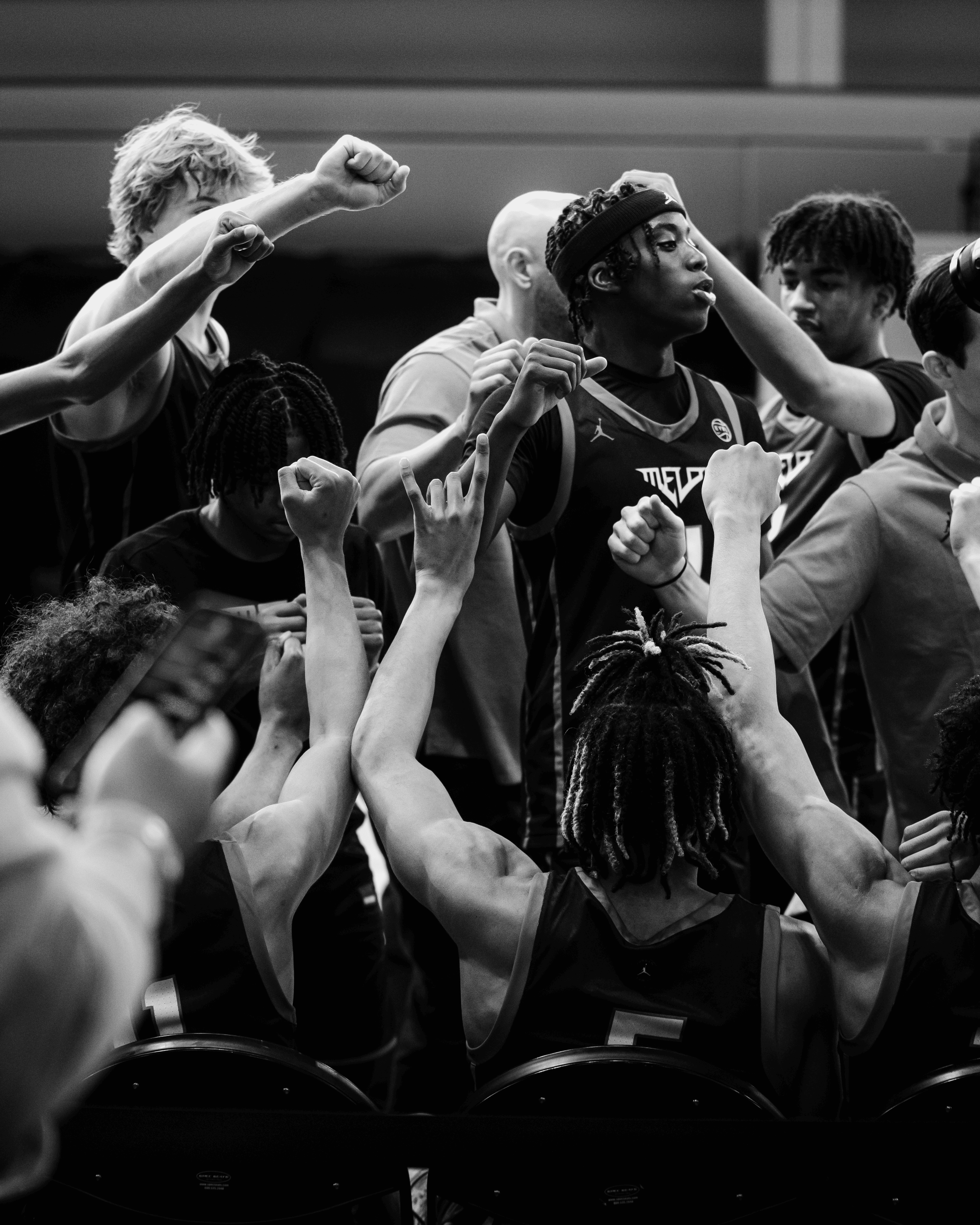 A basketball team huddles together during a timeout or team meeting, with some players raising their fists and arms in solidarity, in a competitive game or practice setting.