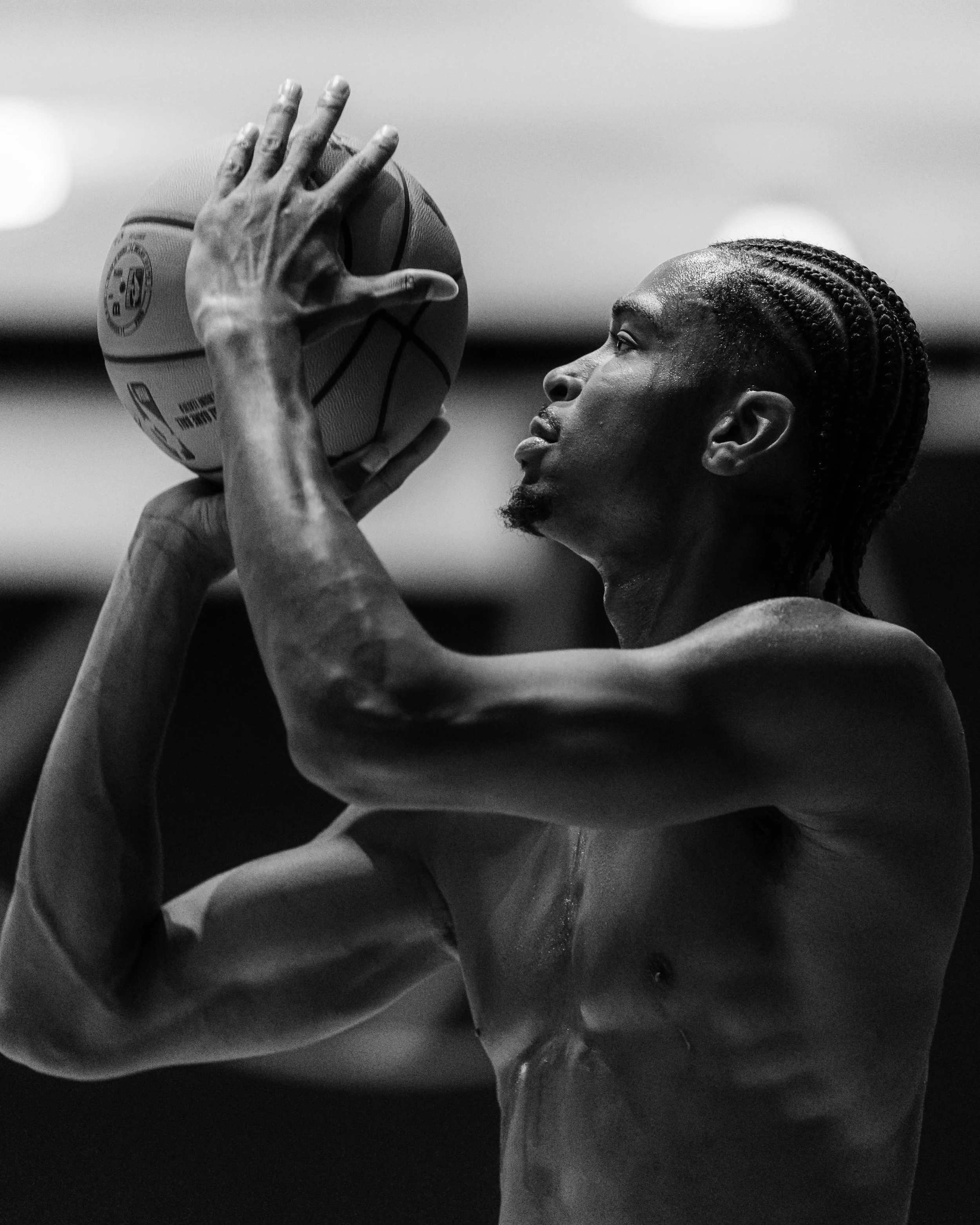 A black and white photo of a shirtless man with braided hair holding a basketball with both hands, preparing to shoot.