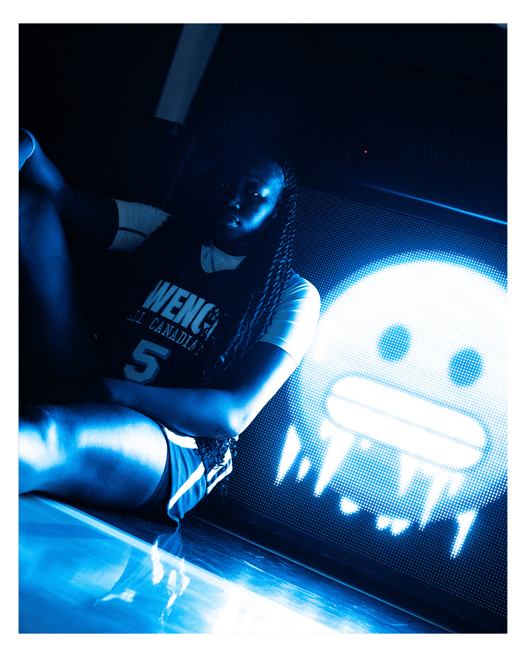 A woman with long braided hair sitting on the floor in a dark room illuminated by a large digital screen displaying a stylized skull with crossbones.