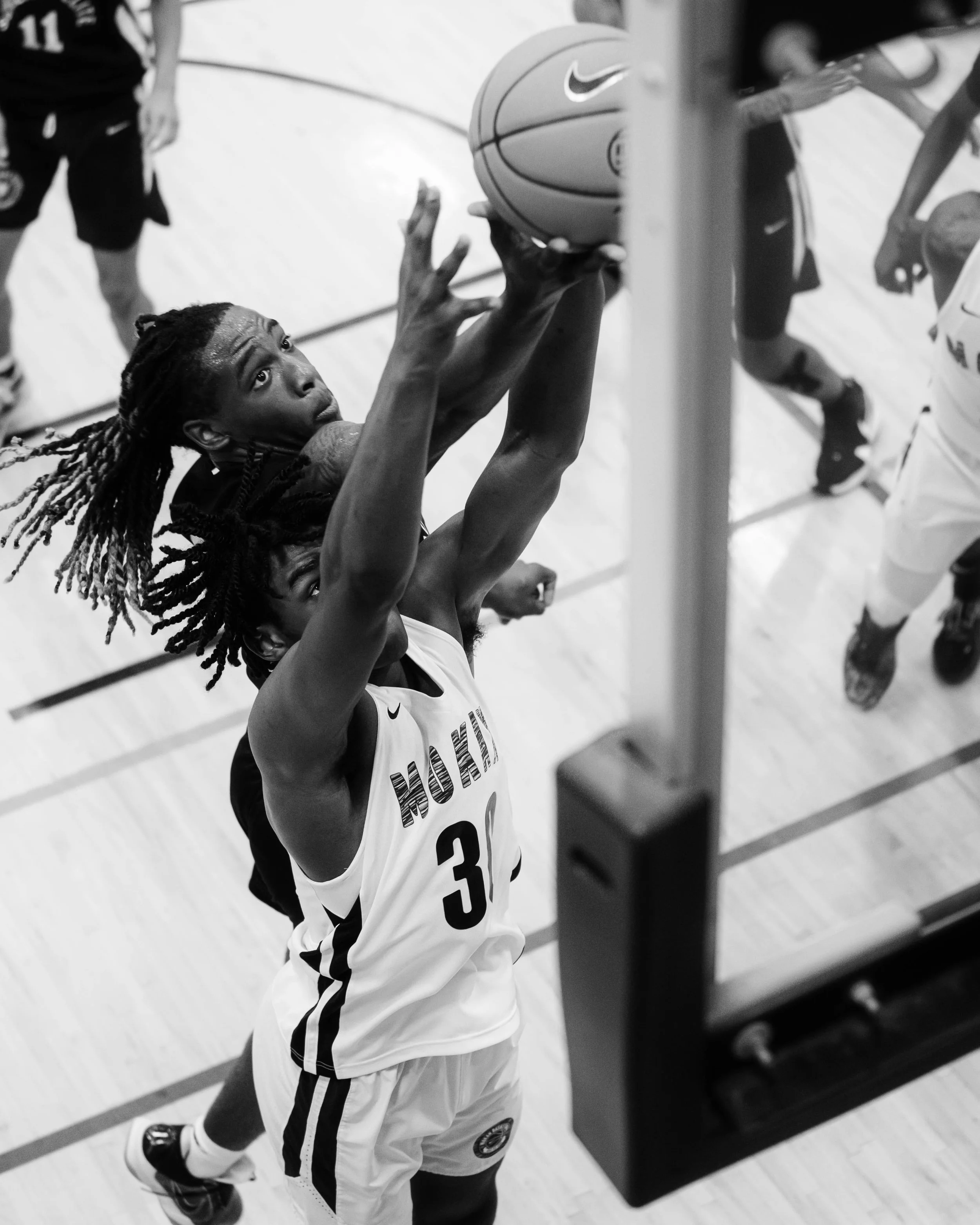 Black and white photo of a basketball player wearing a jersey with the number 3 attempting a shot near the hoop, with other players and a referee visible in the background.