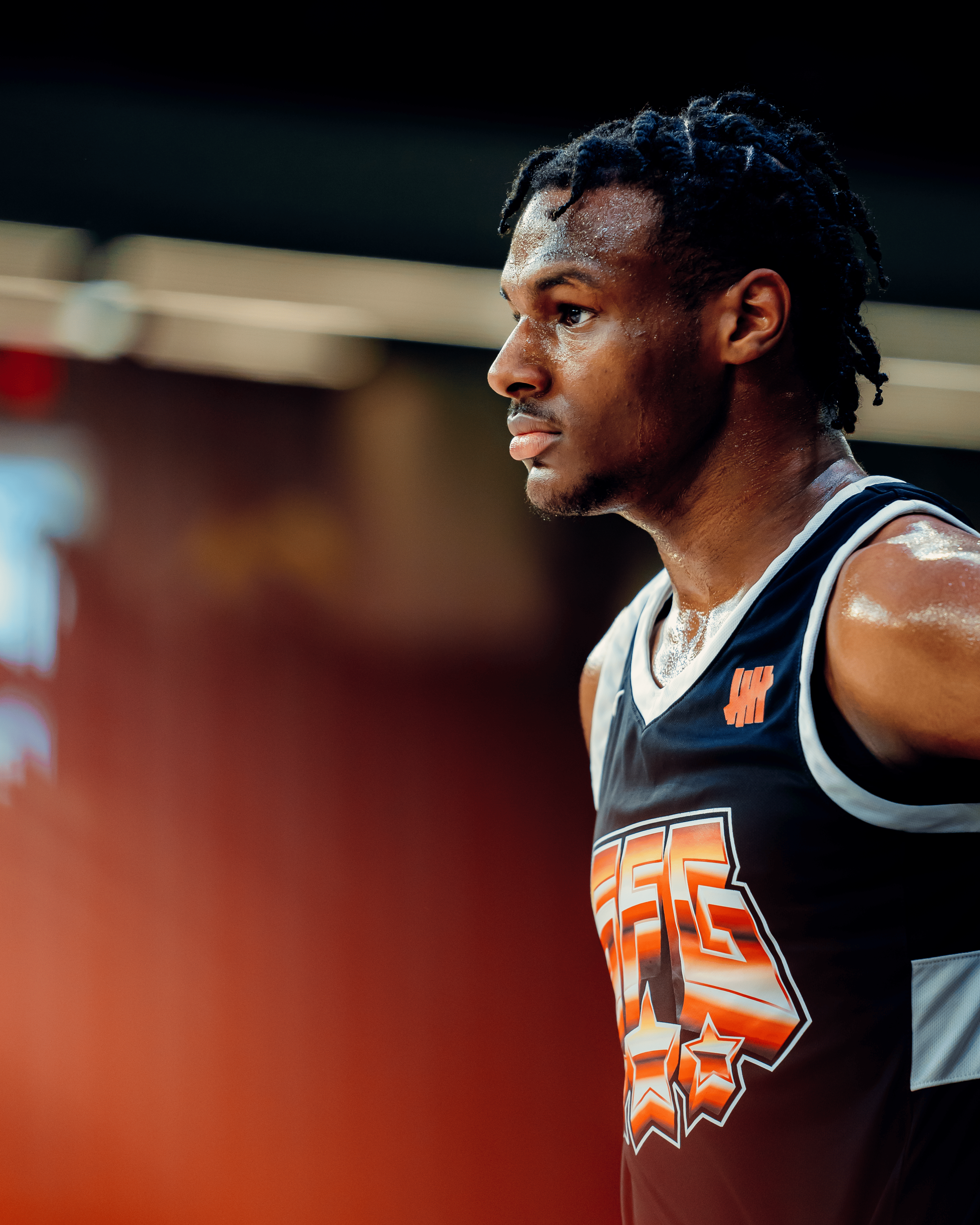 Close-up of a young man in a basketball jersey with sweat, focused and looking to the side, in an indoor gym setting.