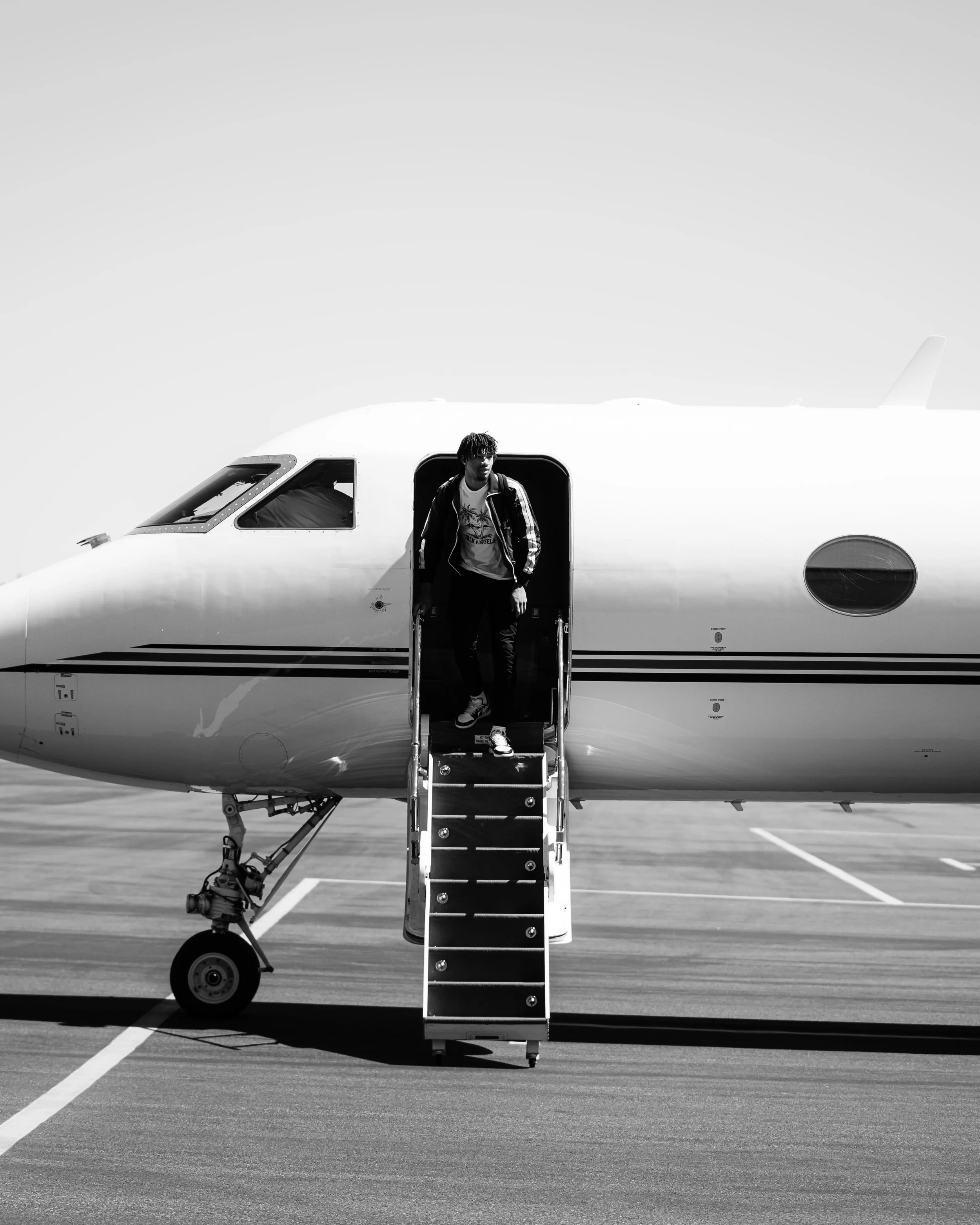 A man in casual clothing in a jacket, t-shirt, and sneakers, is descending the stairs from a private jet on an airport tarmac.