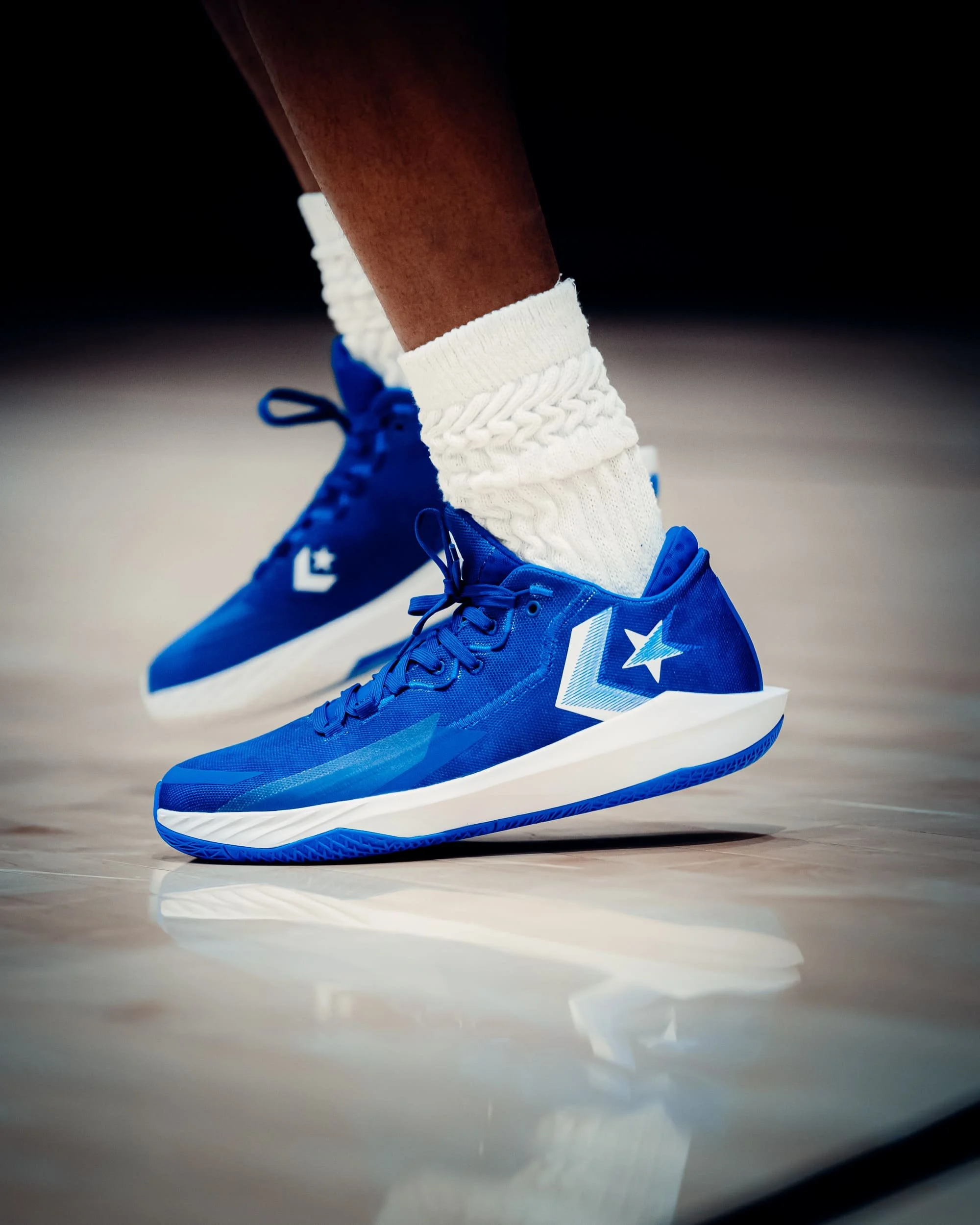 Close-up of a person's feet wearing bright blue athletic sneakers with white soles, paired with white crew socks with cable knit pattern, on a gymnasium or sports court.