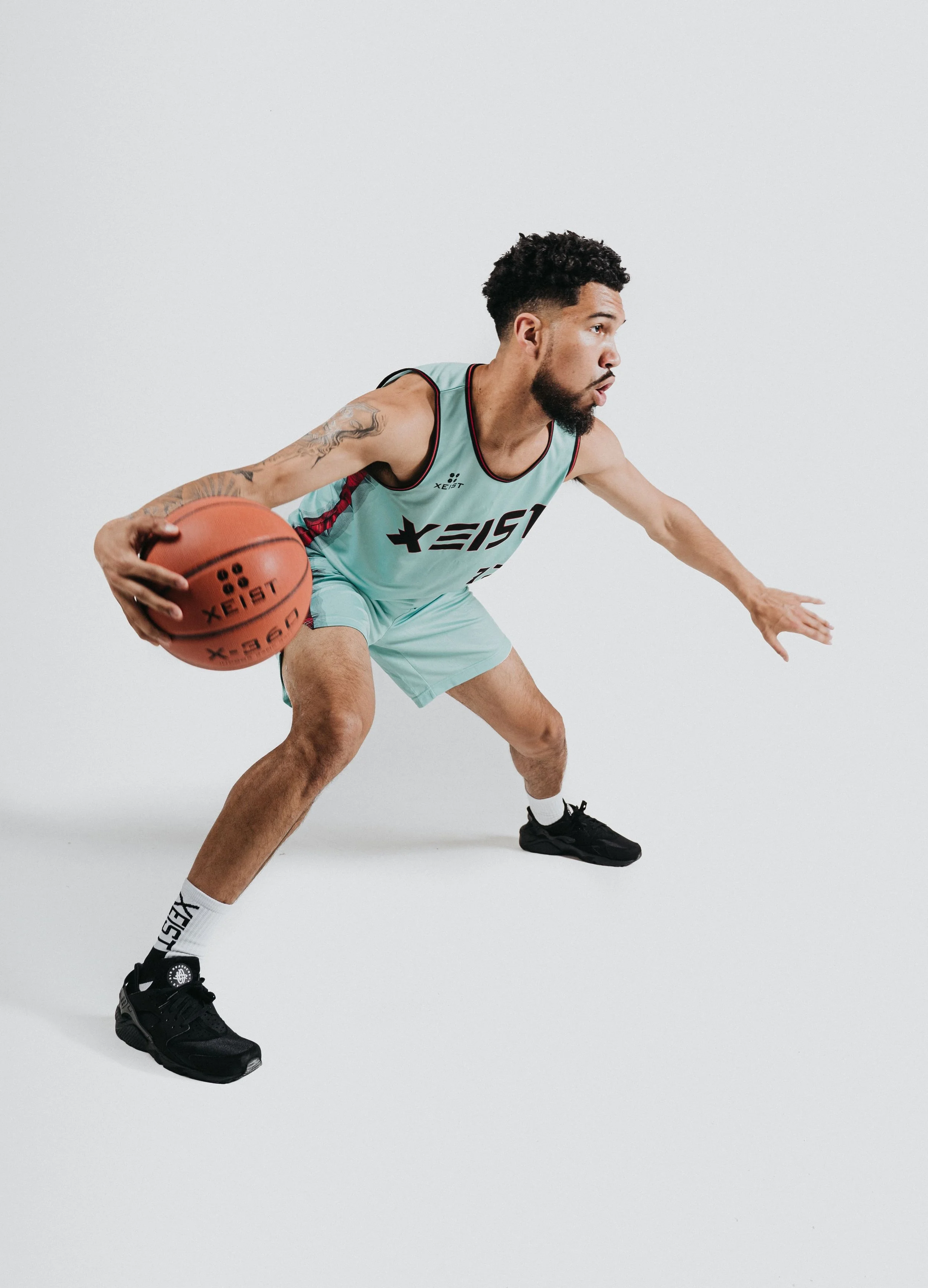 A man wearing a teal basketball uniform dribbling a basketball in a low stance on a plain white background.