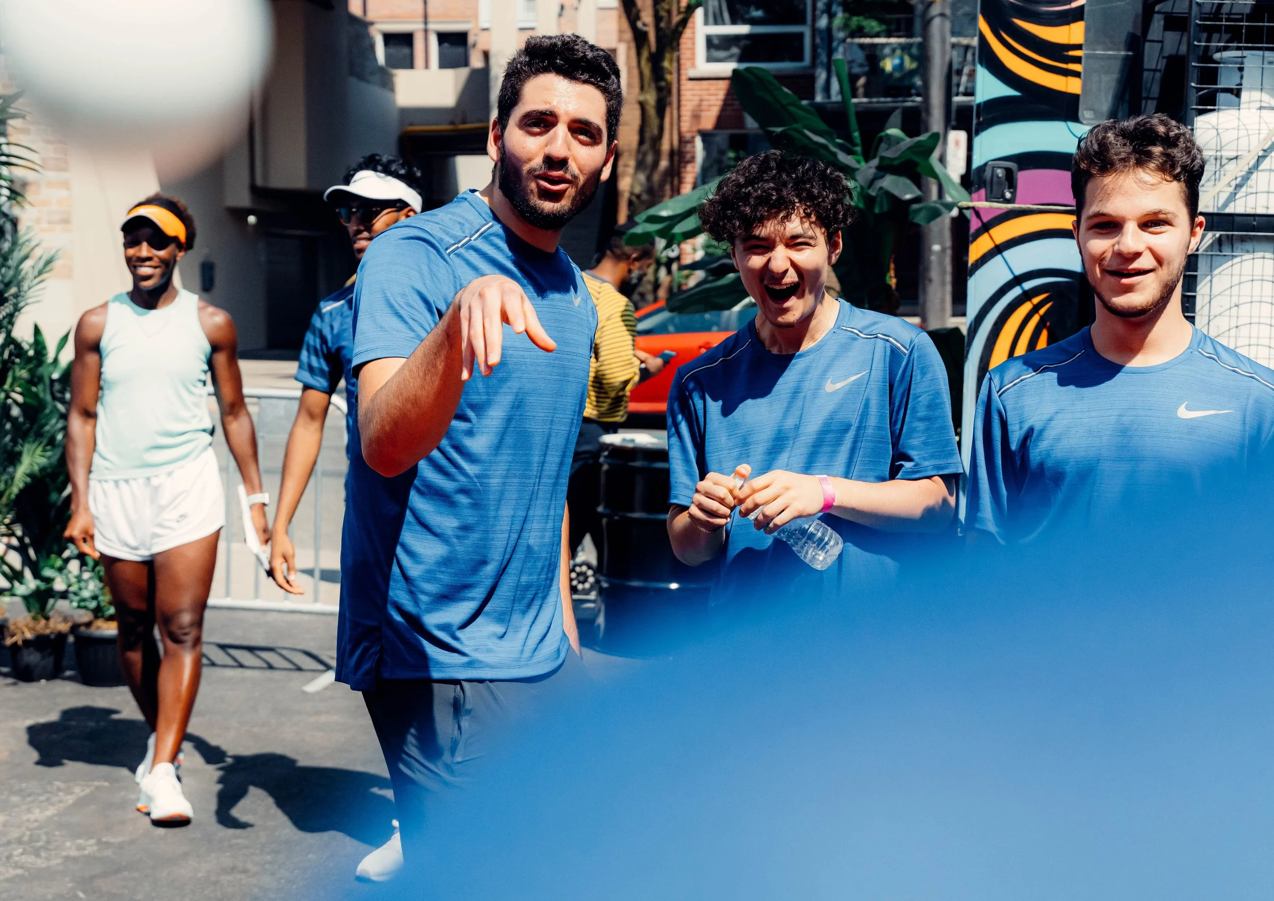 Group of people outdoors, mostly wearing blue Nike athletic shirts, smiling and talking, with a woman in white athletic wear walking behind them, on a sunny day.