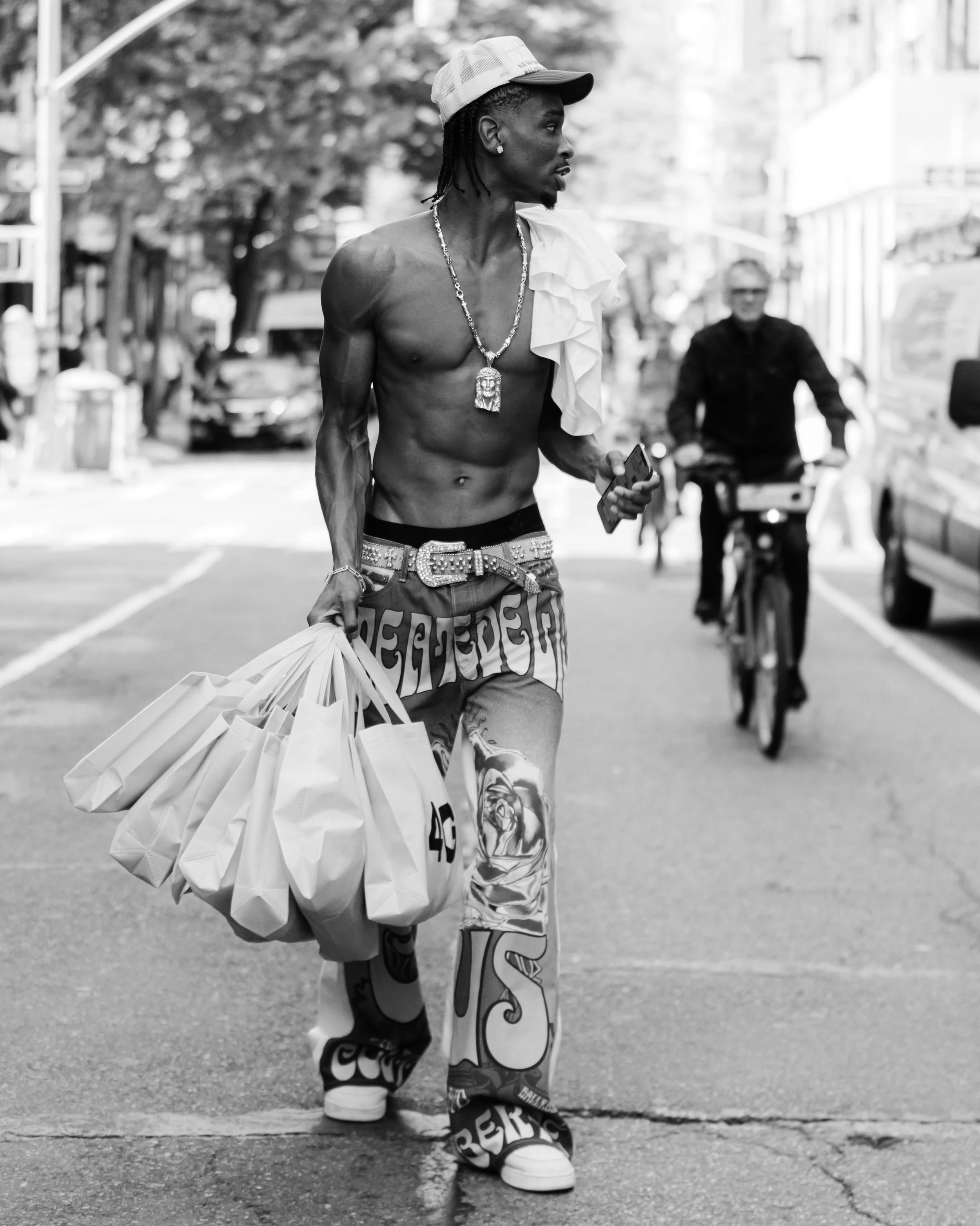 A shirtless man with dreadlocks wearing a cap, jewelry, and patterned pants, walking on the street carrying shopping bags.