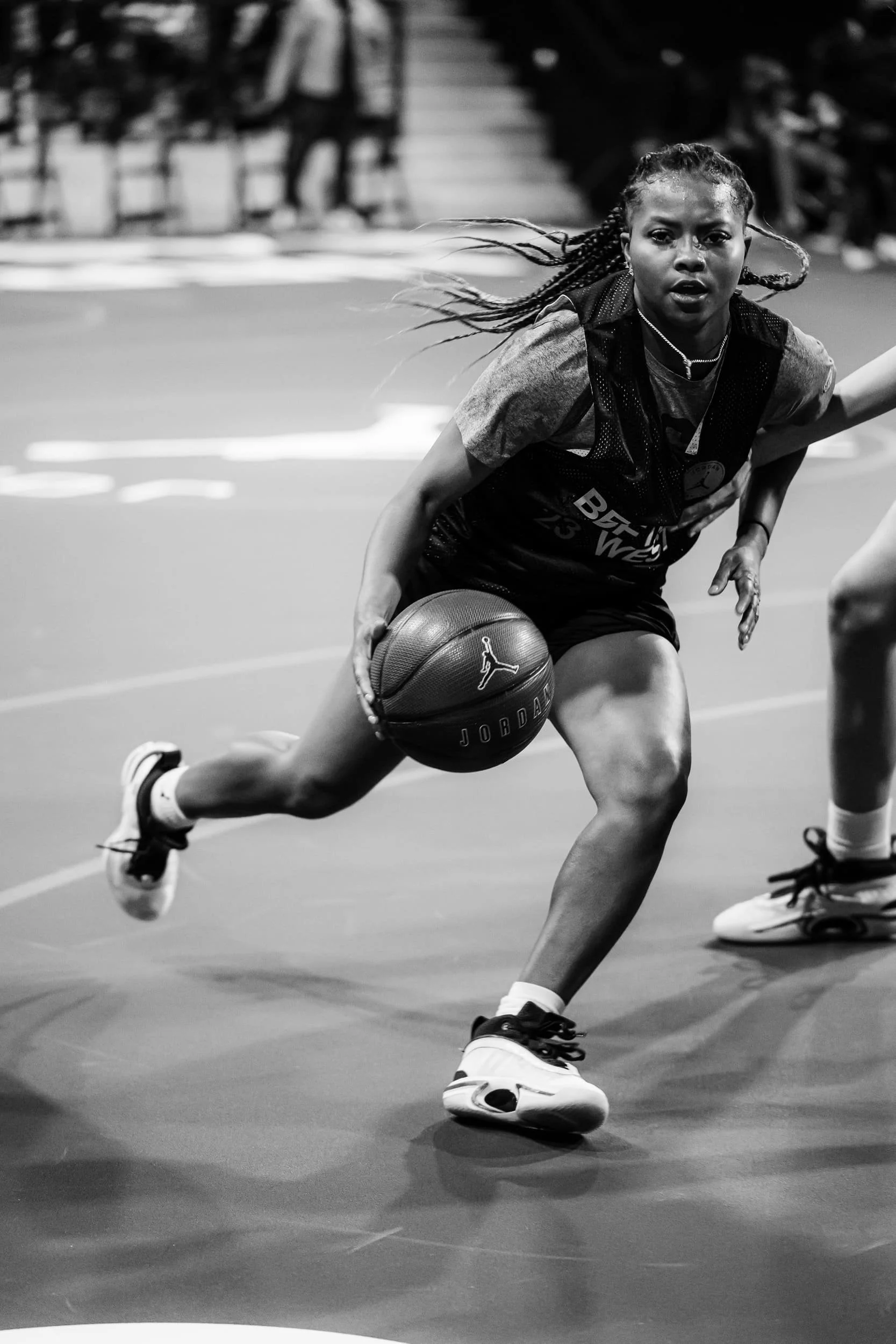 A female basketball player in a black jersey and shorts dribbling a basketball on an indoor court. She has braided hair and is wearing athletic shoes. She appears focused and in motion.