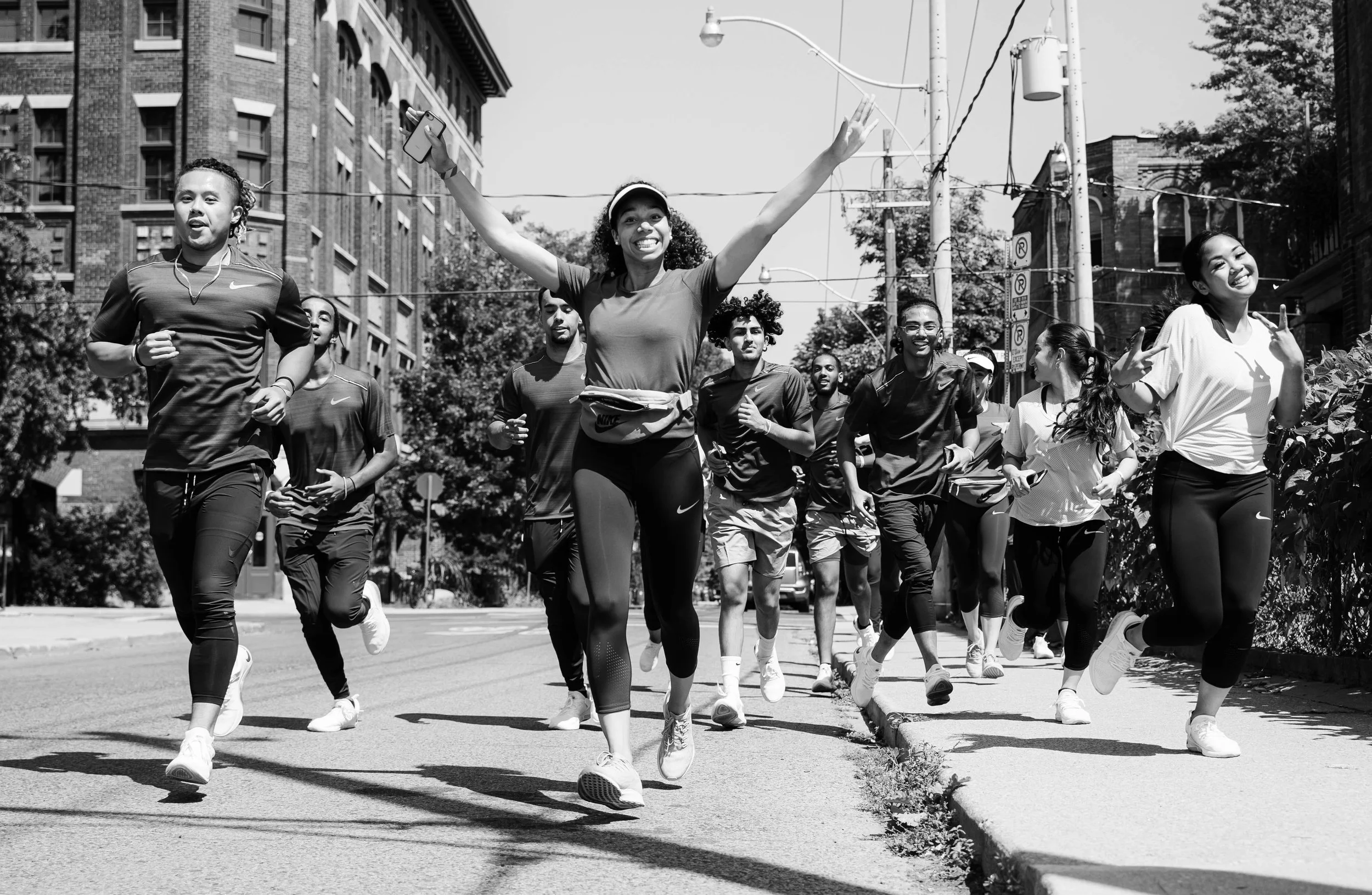 Group of people running together on city street, smiling and enjoying themselves, with an urban background of buildings and trees.