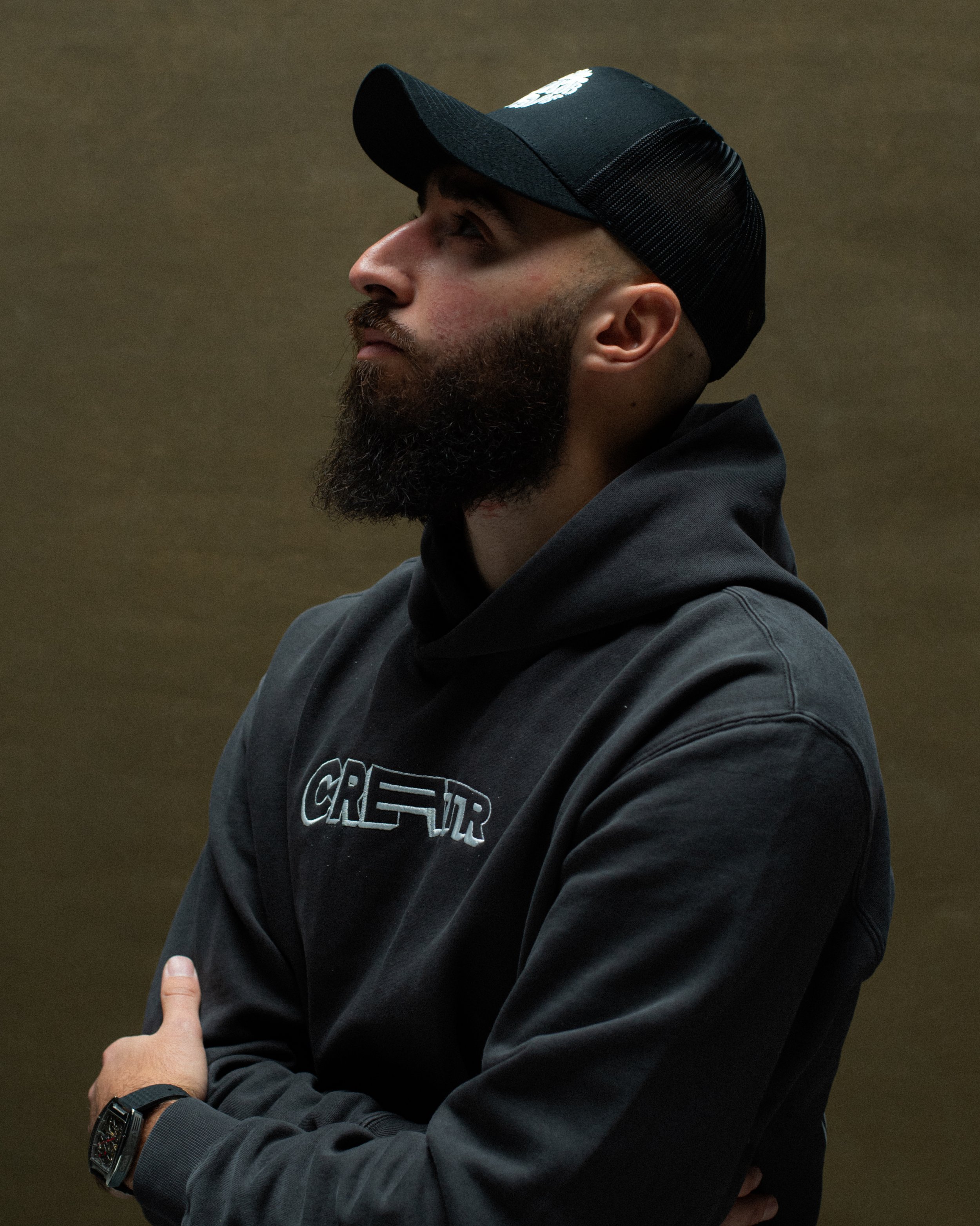 Side profile of a man with a beard wearing a black cap, black hoodie, and wristwatch, standing against a plain background.