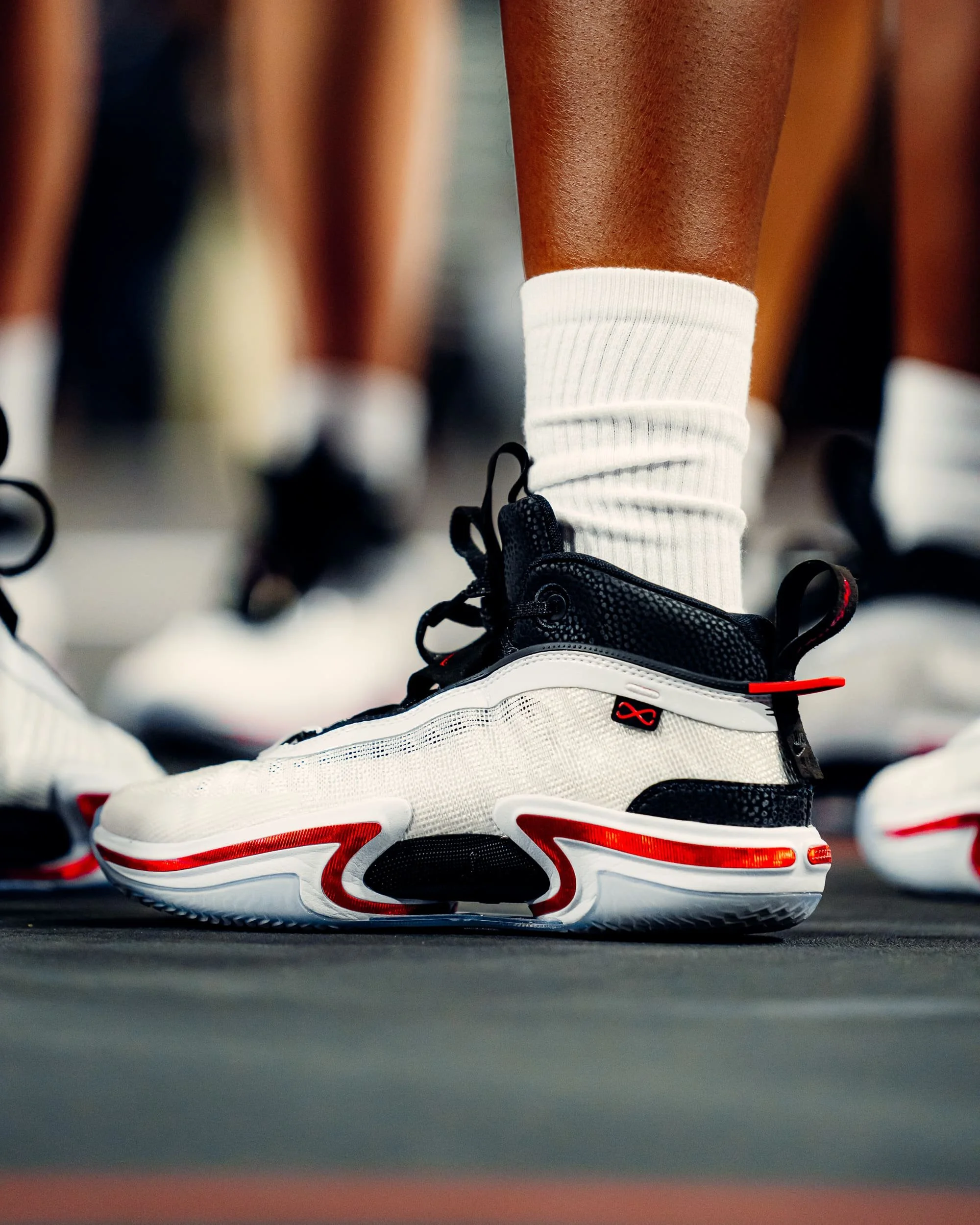 Close-up of a person wearing black and white athletic shoes with red accents, white socks, standing on a gym floor.