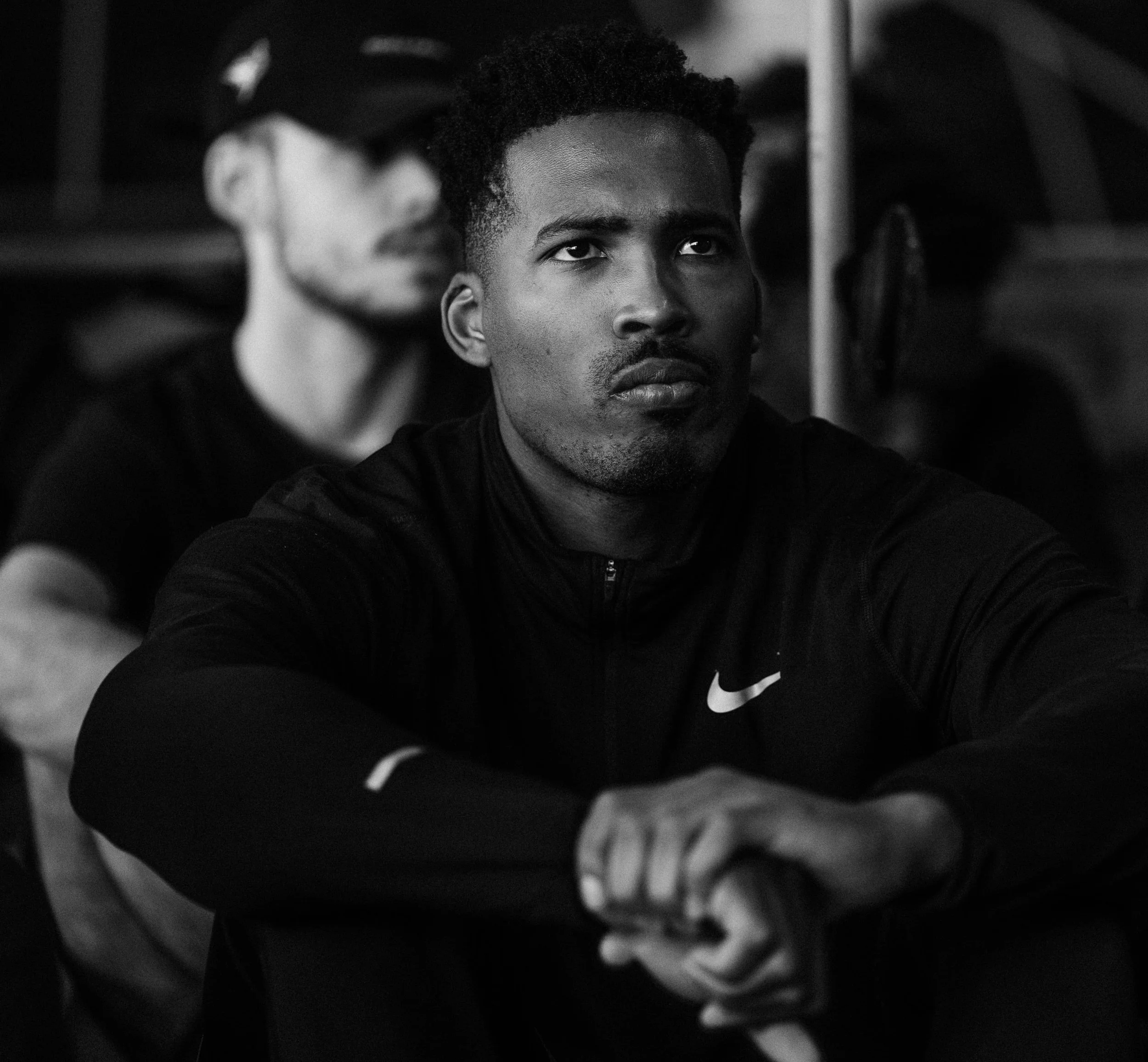 Black and white photo of a young man with short curly hair, wearing a Nike sports jacket, sitting with arms crossed and looking serious. A blurred person wearing glasses in the background.