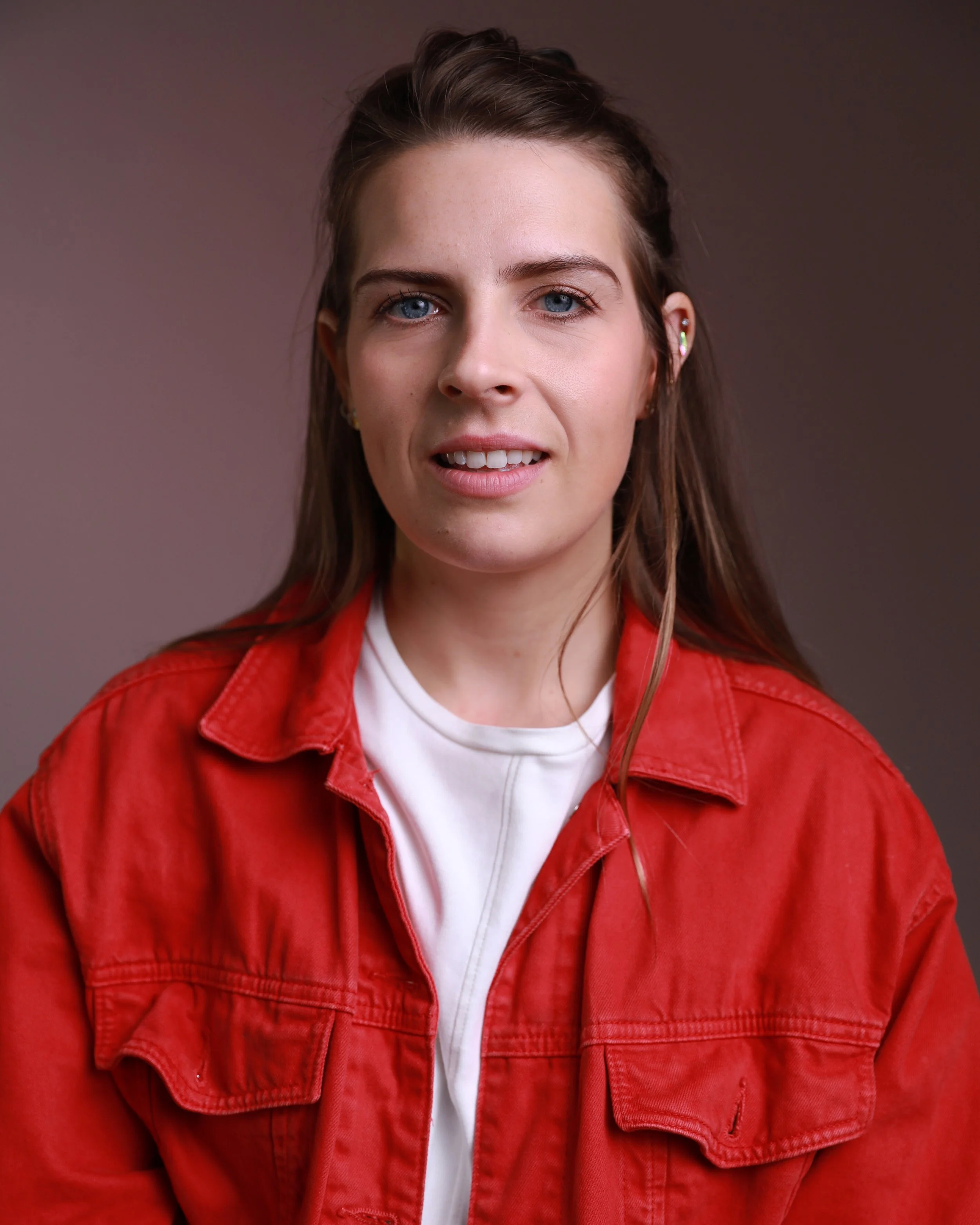 Young woman with blue eyes and brown hair wearing a red jacket over a white shirt, looking at the camera, with a neutral background.