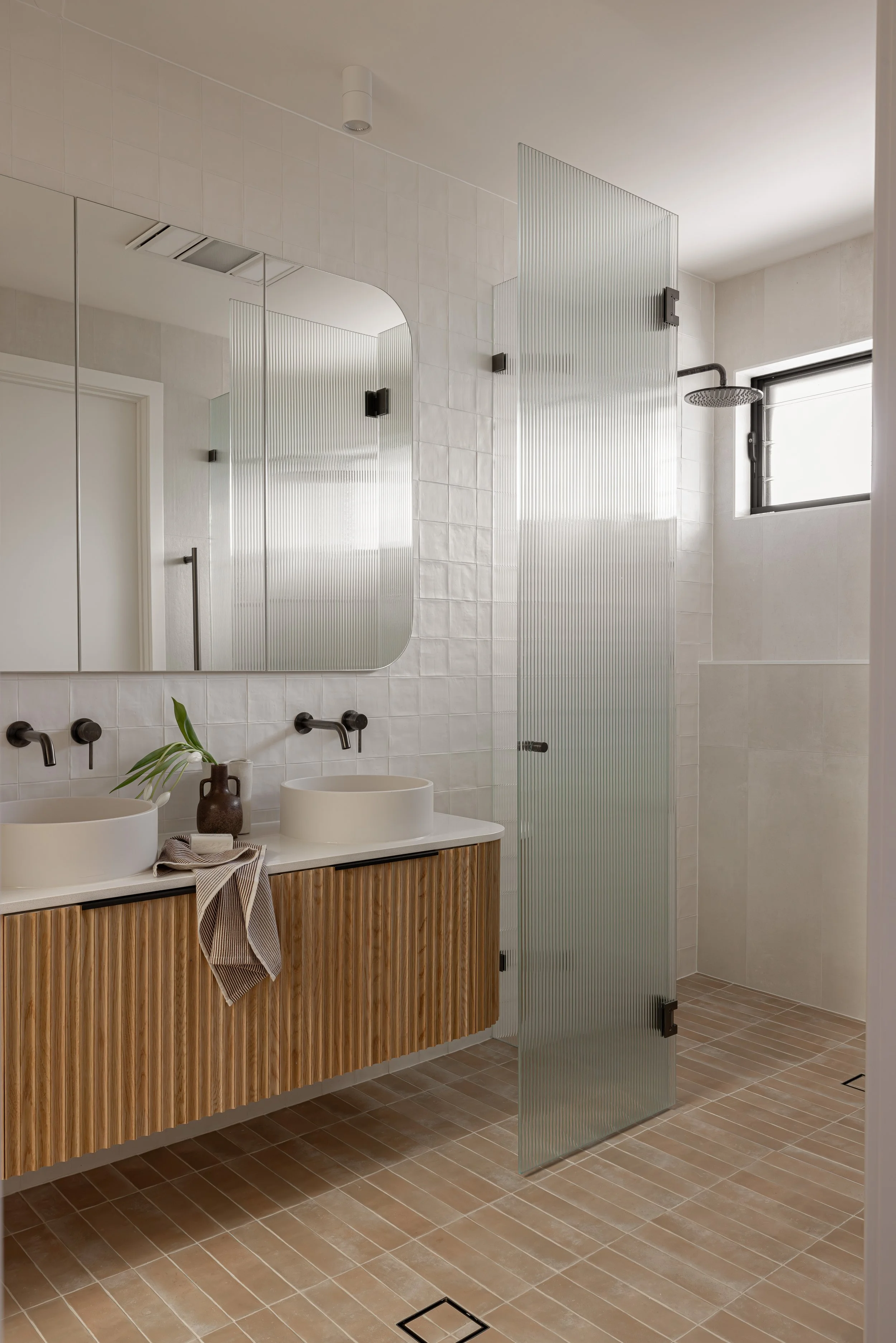 Modern bathroom with double sink vanity, large mirror, and a glass shower partition. Light-colored tiles and a small window provide natural light.