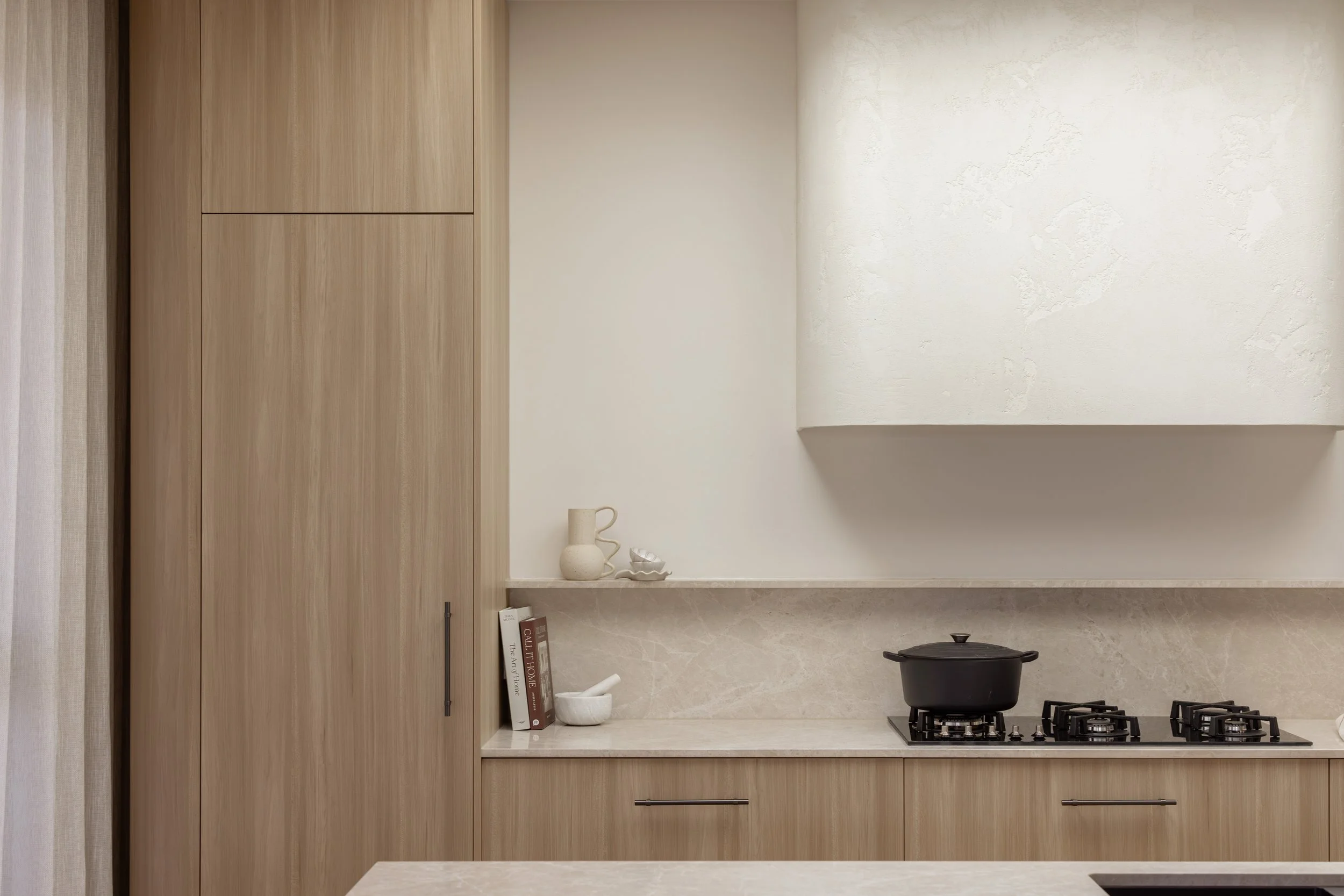 Kitchen with beige marble countertop, black cooking pot on stove, and white ceramic vases and bowls, with wooden cabinets and neutral textured wall
