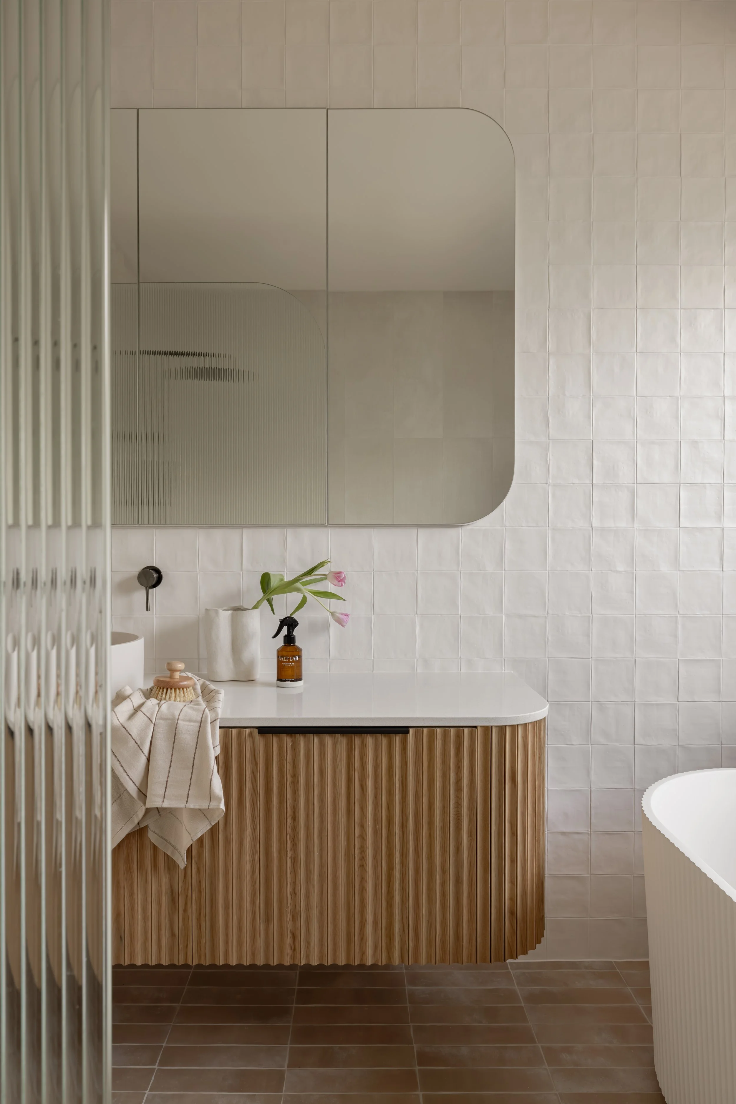 Modern bathroom with a beige tile wall, large mirror above a white countertop, wooden cabinet, and a bathtub partially visible on the right. Decor includes a towel, pink tulips, a soap dispenser, and a dish with a brush.